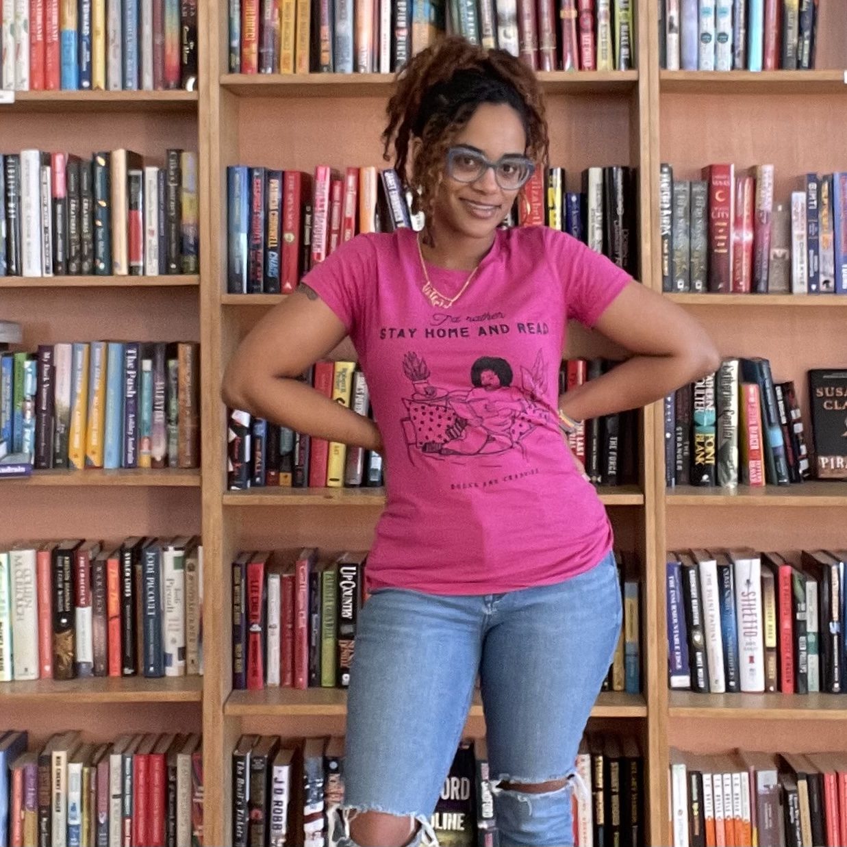 Person stands smiling with hands on hips, wearing a pink shirt and glasses, in front of a large bookshelf filled with assorted books.
