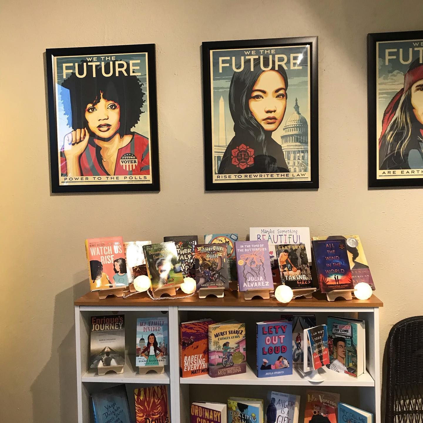Books displayed on a shelf with decorative lighting below framed posters. Posters feature diverse figures with phrases like "Power to the Polls" and "Rewrite the Law."