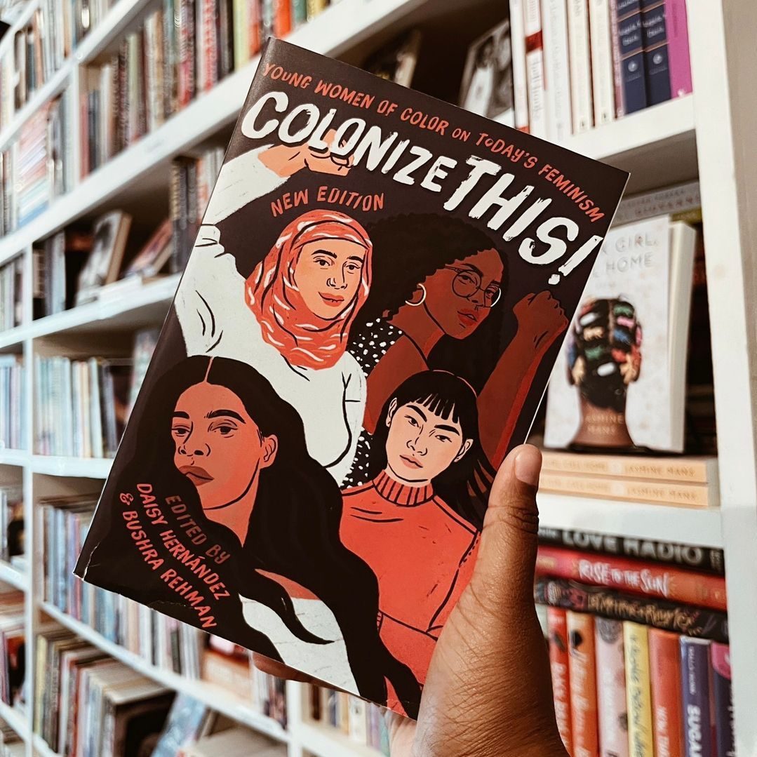 A hand holds a book titled "Colonize This!" featuring an illustration of diverse women, against a backdrop of bookshelves.