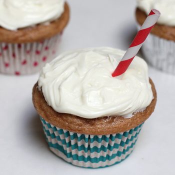 Cupcakes sit on a white surface, topped with frosting and a red-striped straw, surrounded by patterned wrappers.