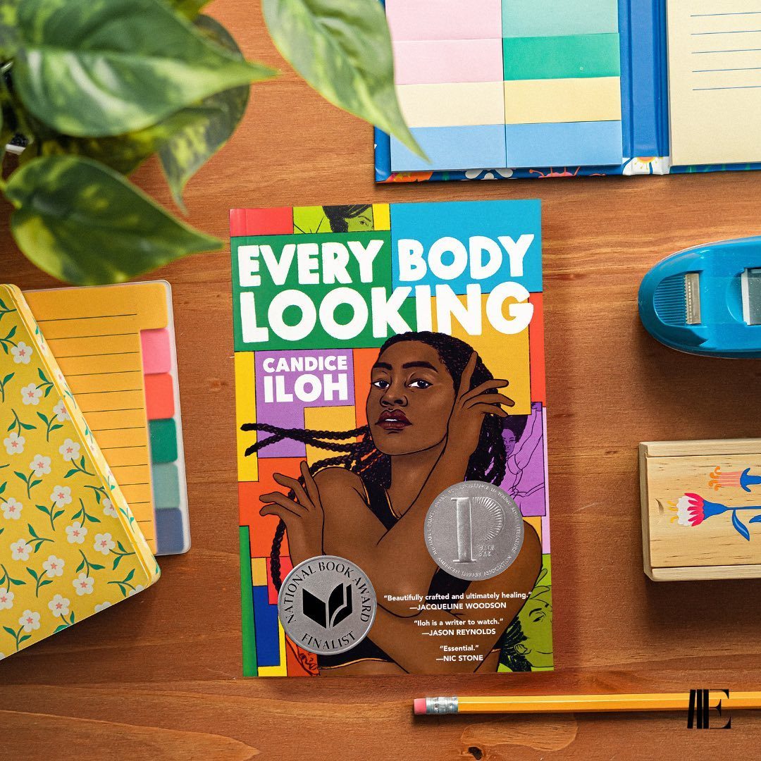 A colorful book titled "Every Body Looking" rests on a wooden desk, surrounded by notebooks, a plant, tape dispenser, and a pencil.