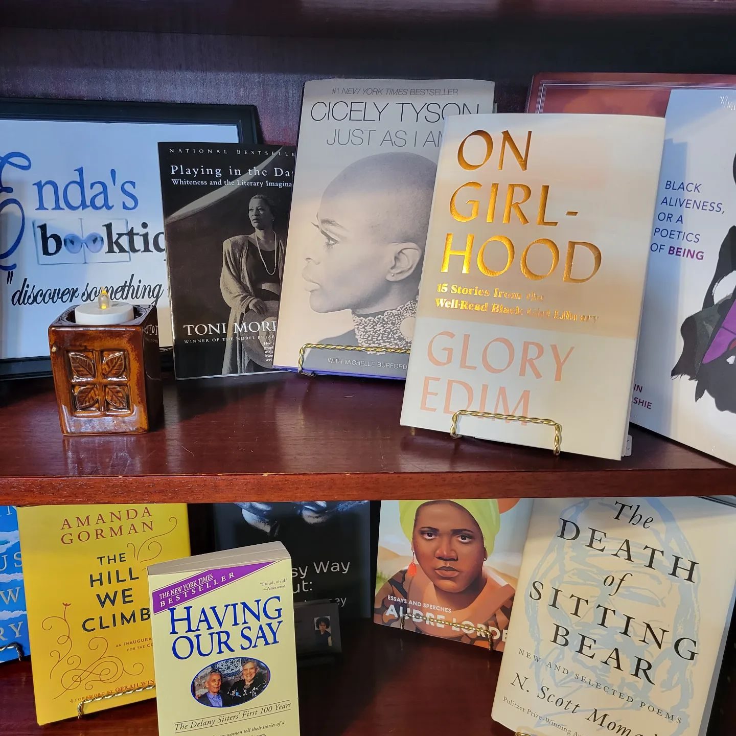 Books rest on wooden shelves, displaying titles like "On Girlhood" by Glory Edim. A small decorative item sits near a bookstore sign.