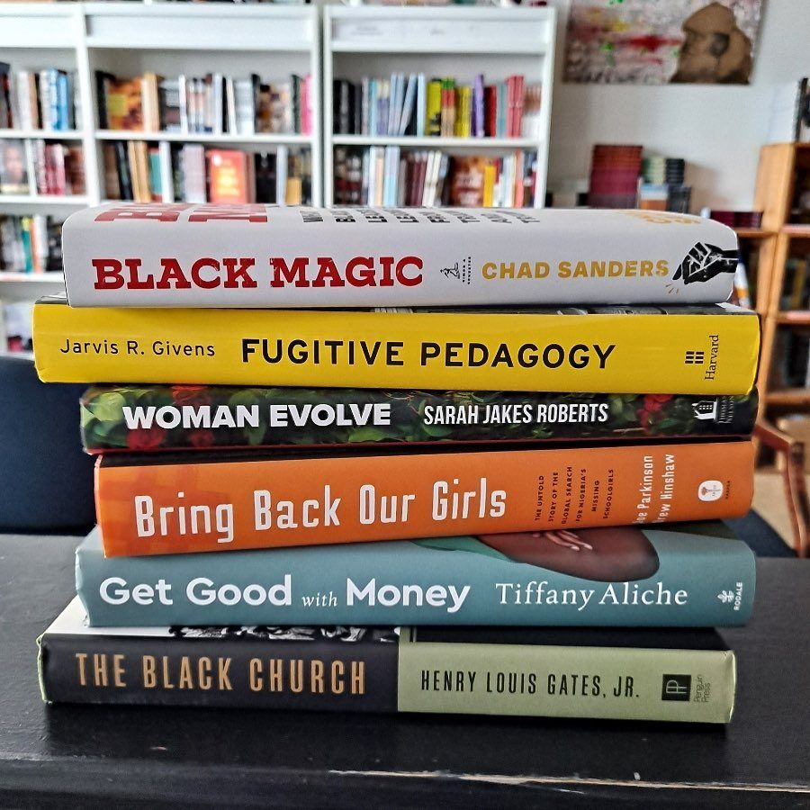 Stack of six books resting on a table, situated in front of a full bookshelf in a room filled with books and shelves.