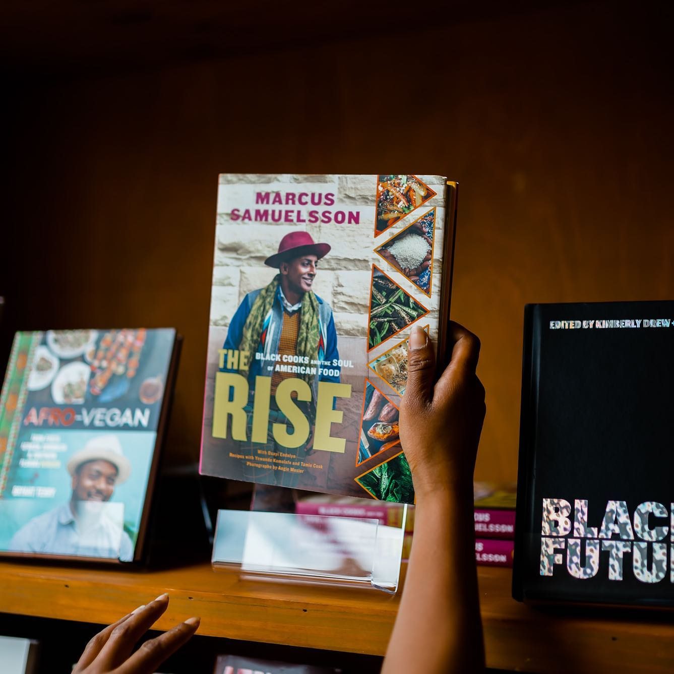 Hands hold a cookbook titled "The Rise" by Marcus Samuelsson on a wooden shelf among other culinary books.