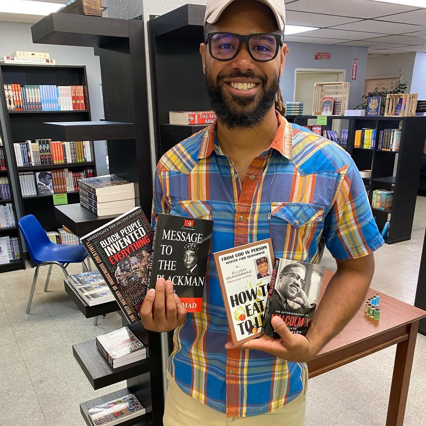 Man holding books, smiling, in a bookstore. Books include "Black People Invented Everything," "Message to the Blackman," "How to Eat to Live," and "The Autobiography of Malcolm X."
