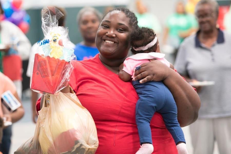 A smiling woman holds a baby and a gift bag in a crowded, festive setting with balloons.