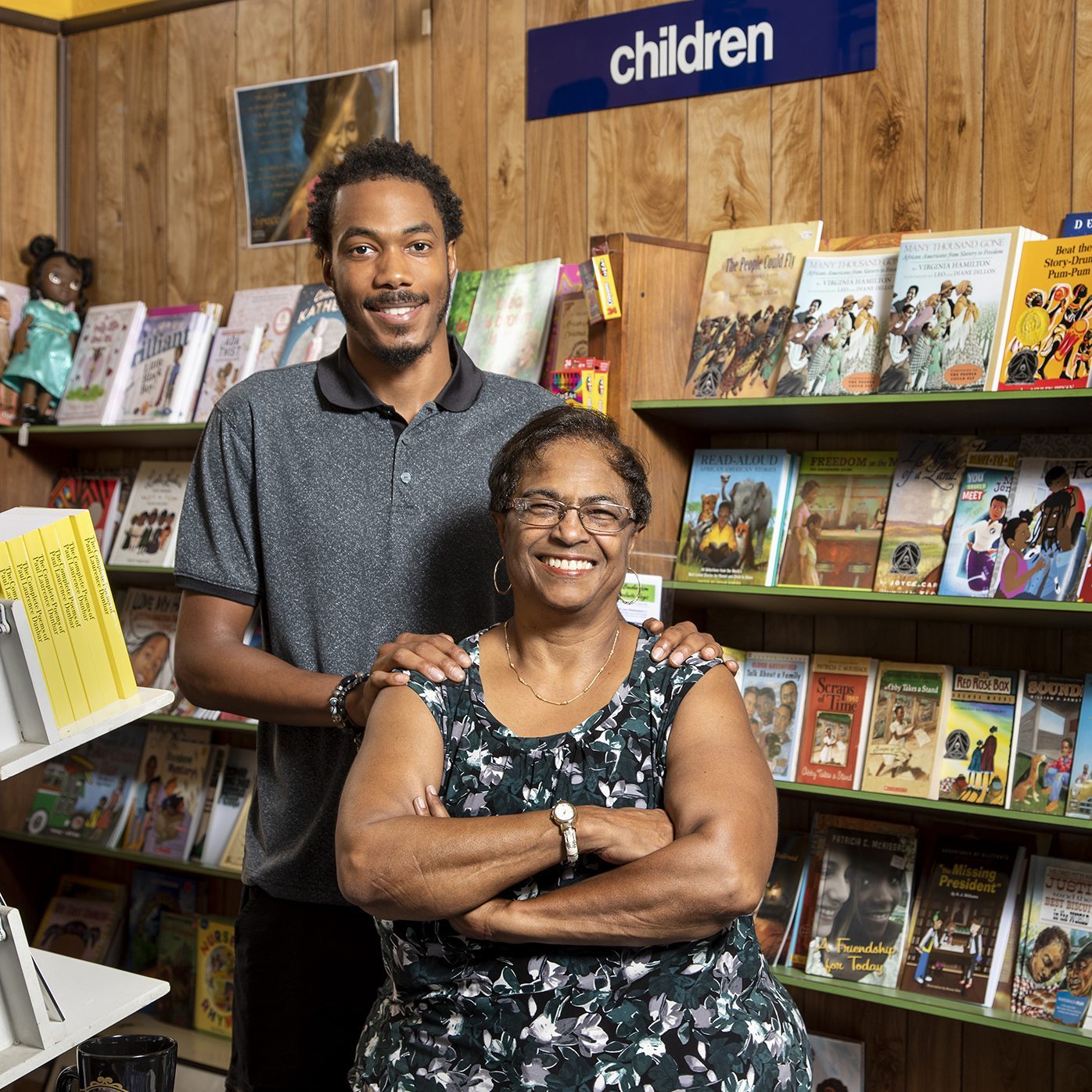 Two people smile, standing in a bookstore's children's section, surrounded by shelves filled with colorful children's books.
