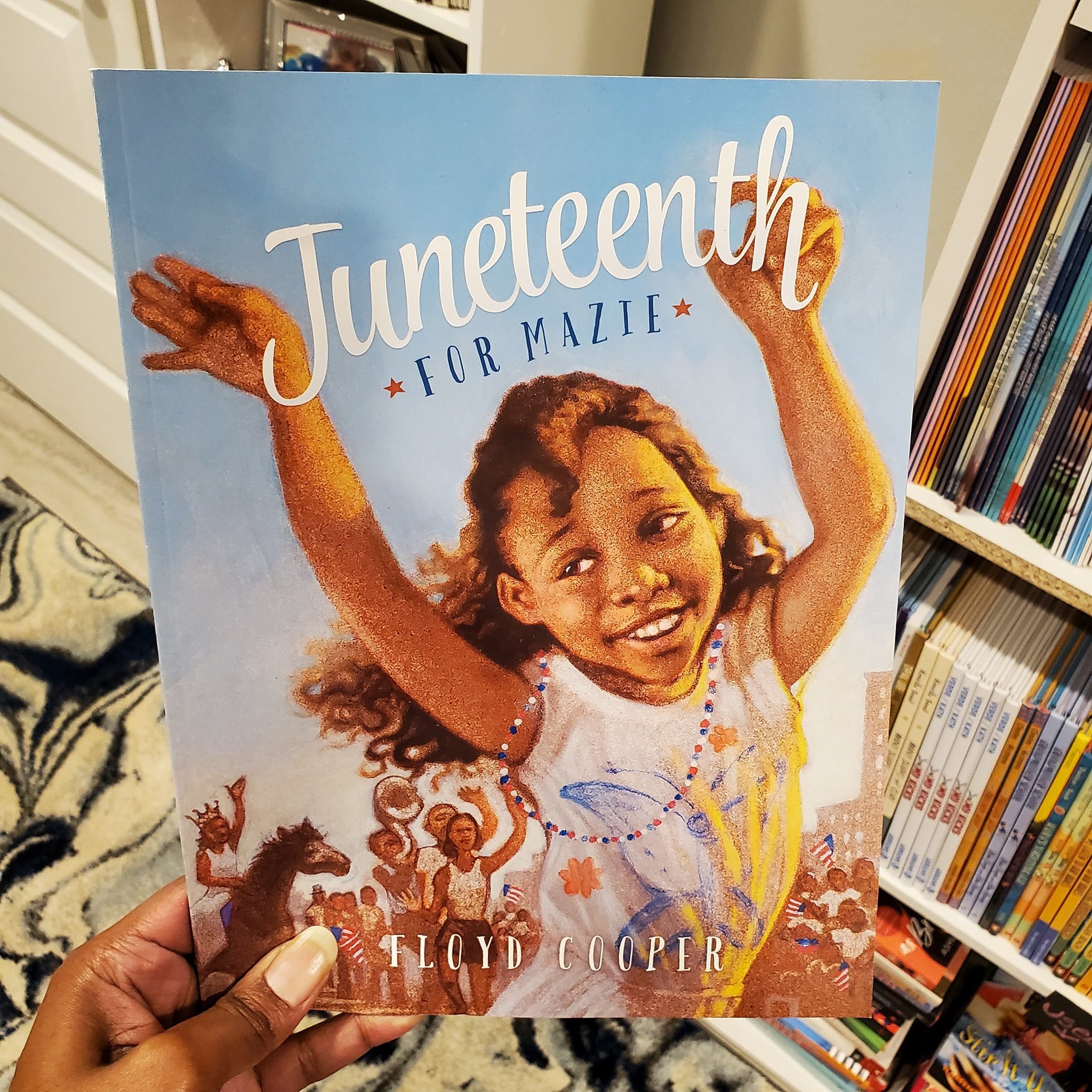 Book cover depicts a smiling child raising arms in celebration, surrounded by joyful people, indoors near shelves with books. Title: "Juneteenth for Mazie" by Floyd Cooper.