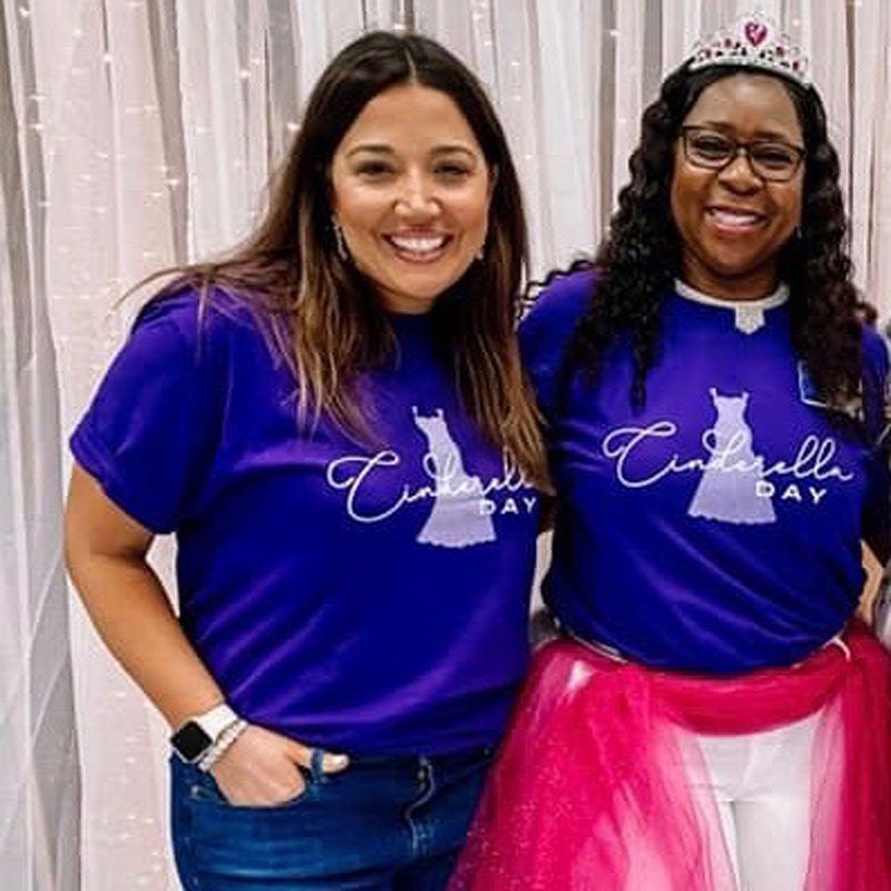 Two smiling women wear "Cinderella Day" t-shirts; one has a tiara. They stand in front of a light-colored curtain backdrop.