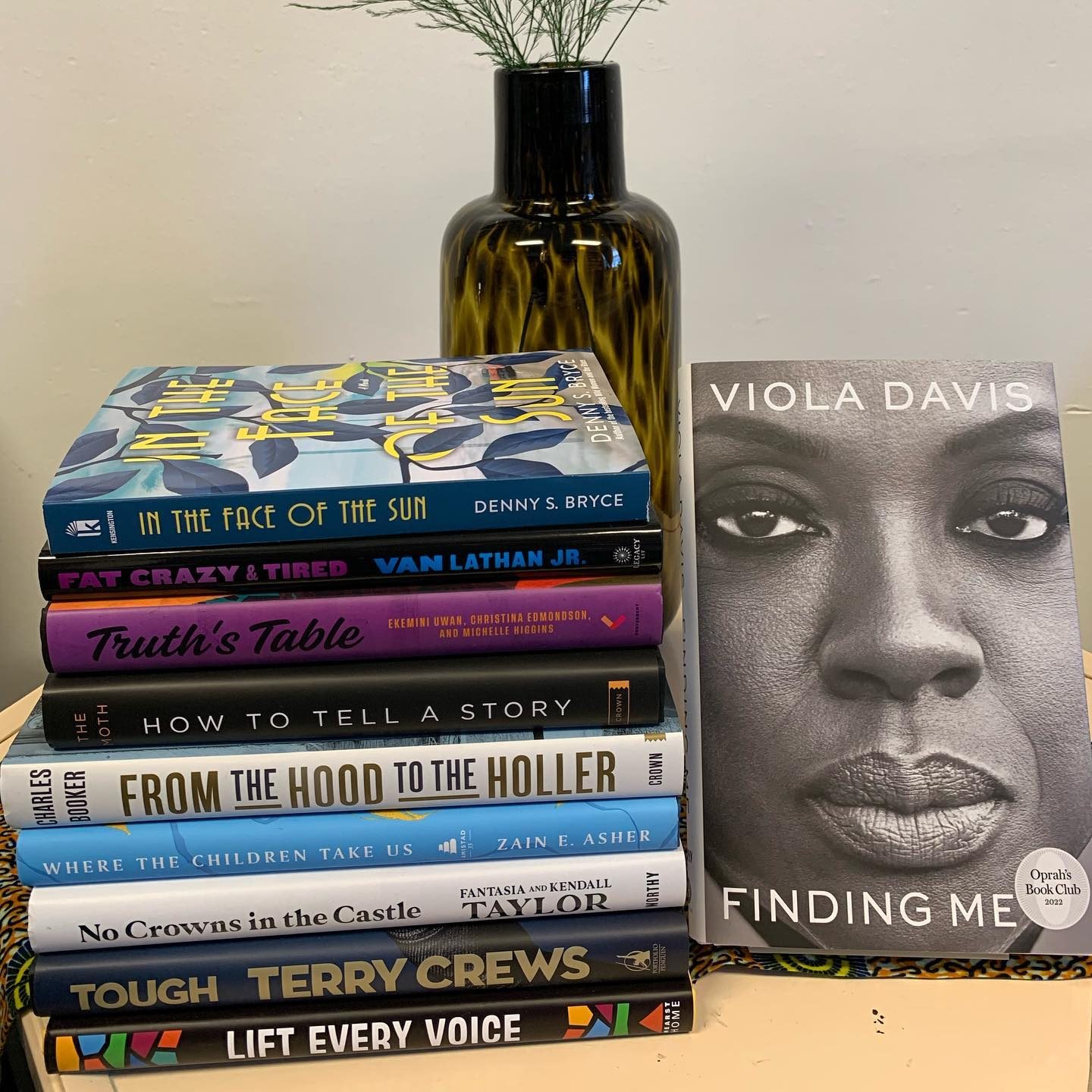 Books are stacked with "Finding Me" by Viola Davis upright. A dark vase with green stems sits nearby on a light-colored surface.