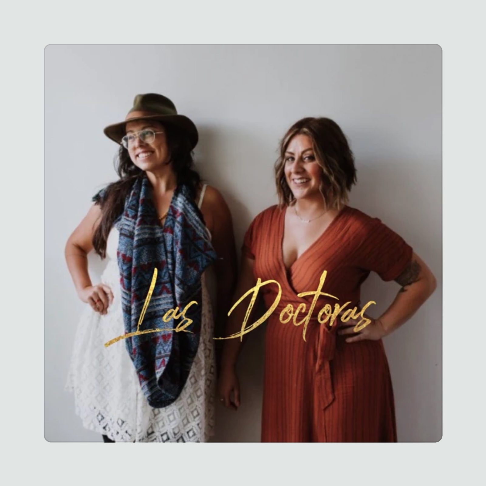 Two women stand smiling against a plain background, wearing casual dresses and accessories. Text reads, "Las Doctoras."