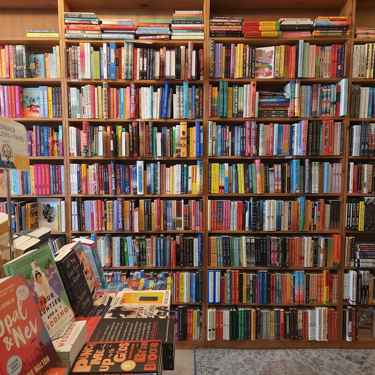 Books fill wooden shelves; some are displayed upright on a table. A sign reads "Paperback Reader Faves." The room has a cozy atmosphere.