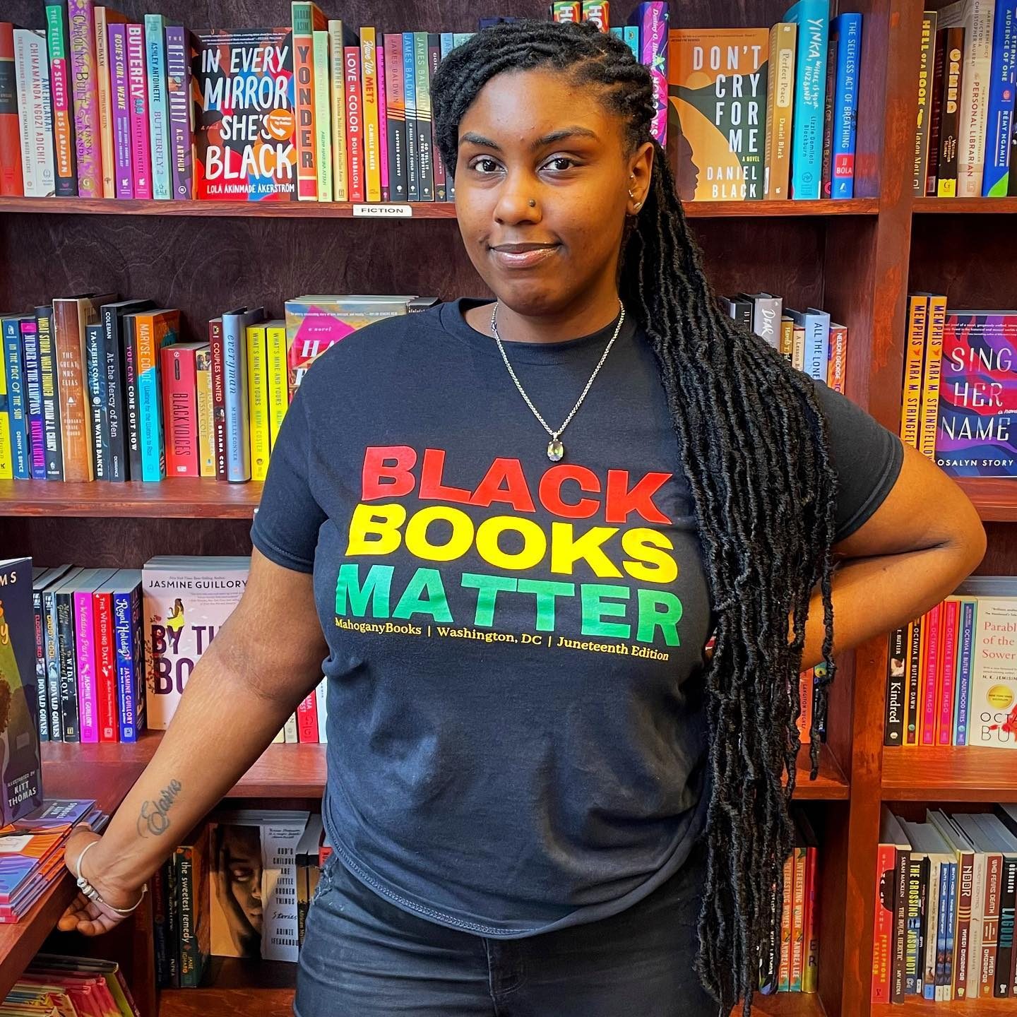 Person stands confidently, wearing "BLACK BOOKS MATTER" shirt; surrounded by colorful books on wooden shelves, creating a bookstore environment.