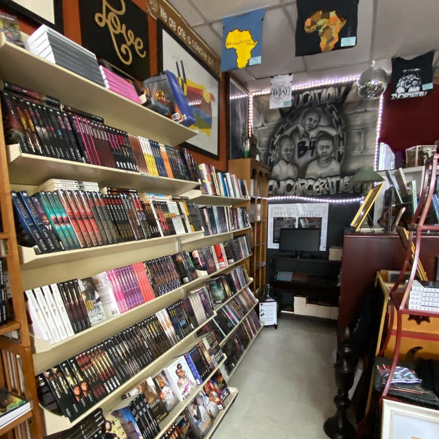 Books fill shelves; art and flags decorate the vibrant store. Text reads â€œEducation B4 Incarcerationâ€ and "Love." The atmosphere is lively and diverse.