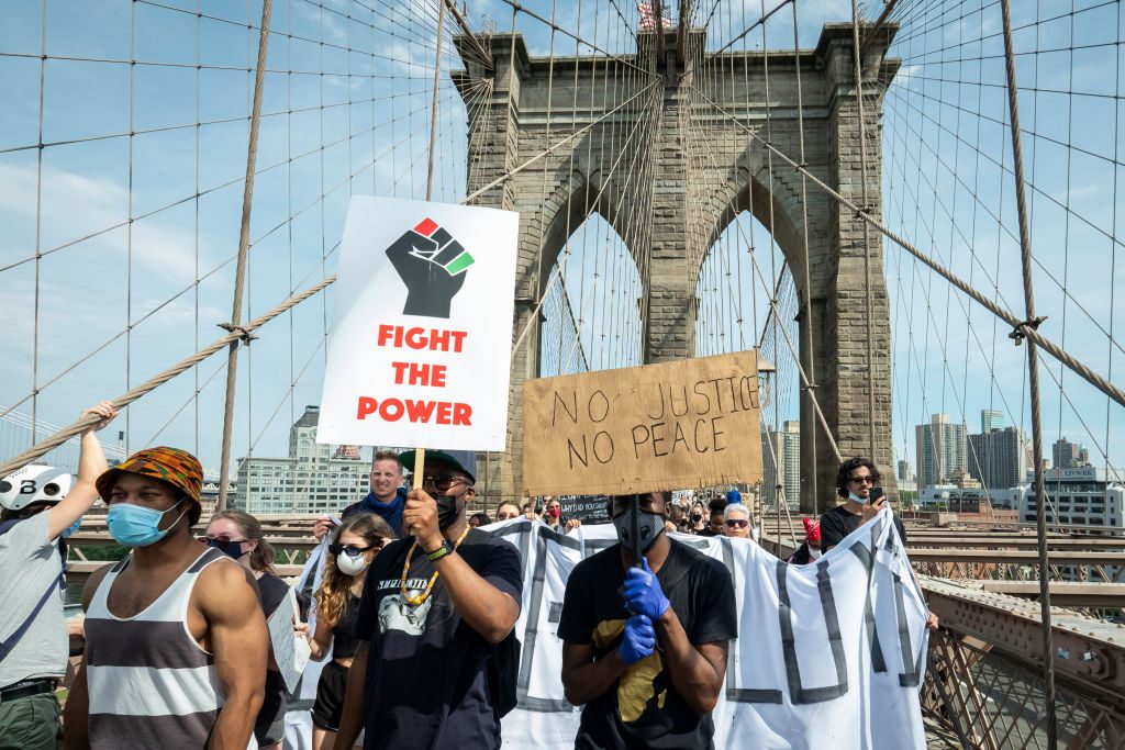 Protesters walk across a bridge holding signs; one reads "FIGHT THE POWER" and another "NO JUSTICE NO PEACE." The city skyline is visible.