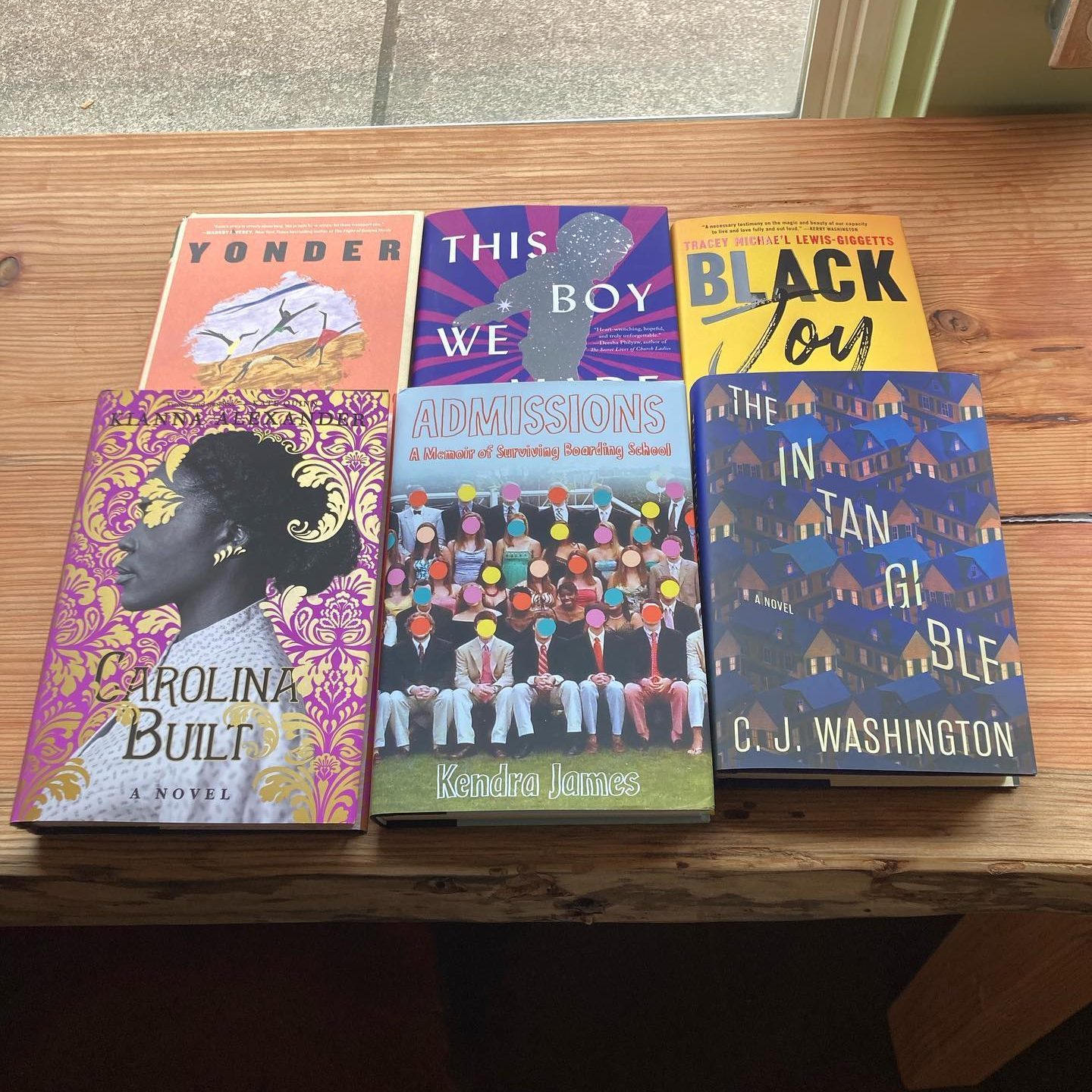 Books are arranged on a wooden table, showcasing colorful covers with titles: "Yonder," "This Boy We Made," "Black Joy," "Carolina Built," "Admissions," "The Intangible."