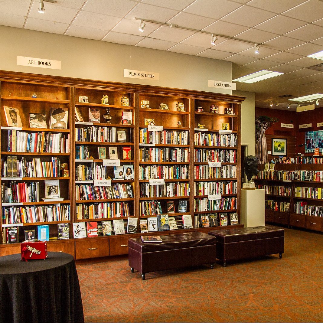 Books fill wooden shelves labeled "Art Books," "Black Studies," and "Biographies" in a bookstore. Benches and a table are in front.
