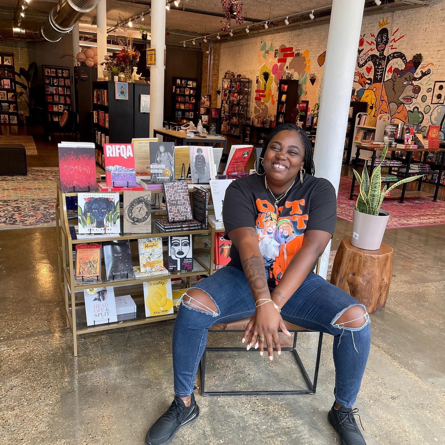 A person smiles while sitting on a chair in a vibrant bookstore, surrounded by books and colorful wall art.