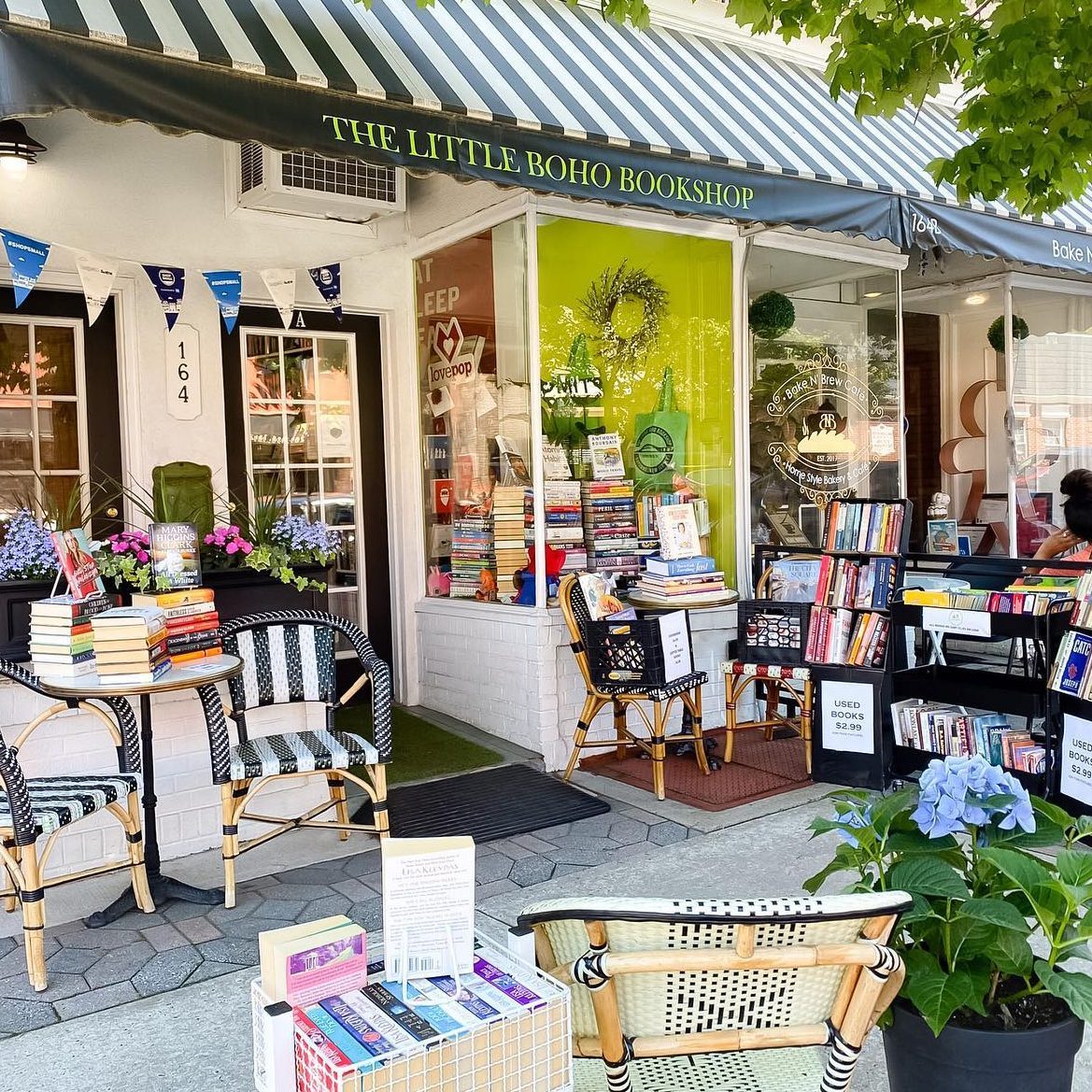 Bookshop displays books on tables and shelves outside. Chairs and potted plants create a cozy, inviting street scene. Sign: "Used Books $2.99."