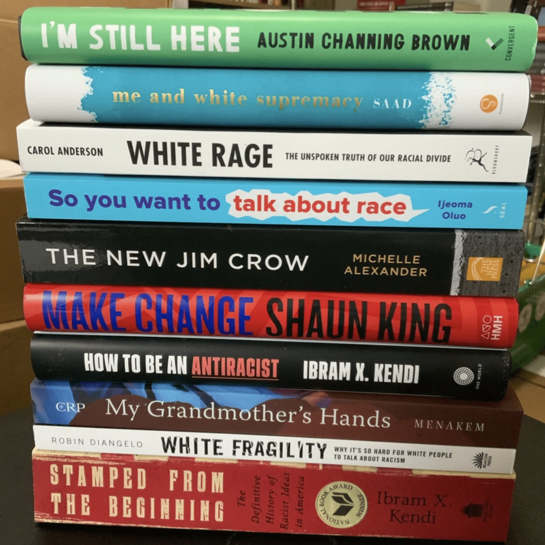 Books stacked horizontally, showing titles addressing race and social justice themes, placed on a wooden surface in a well-lit room.
