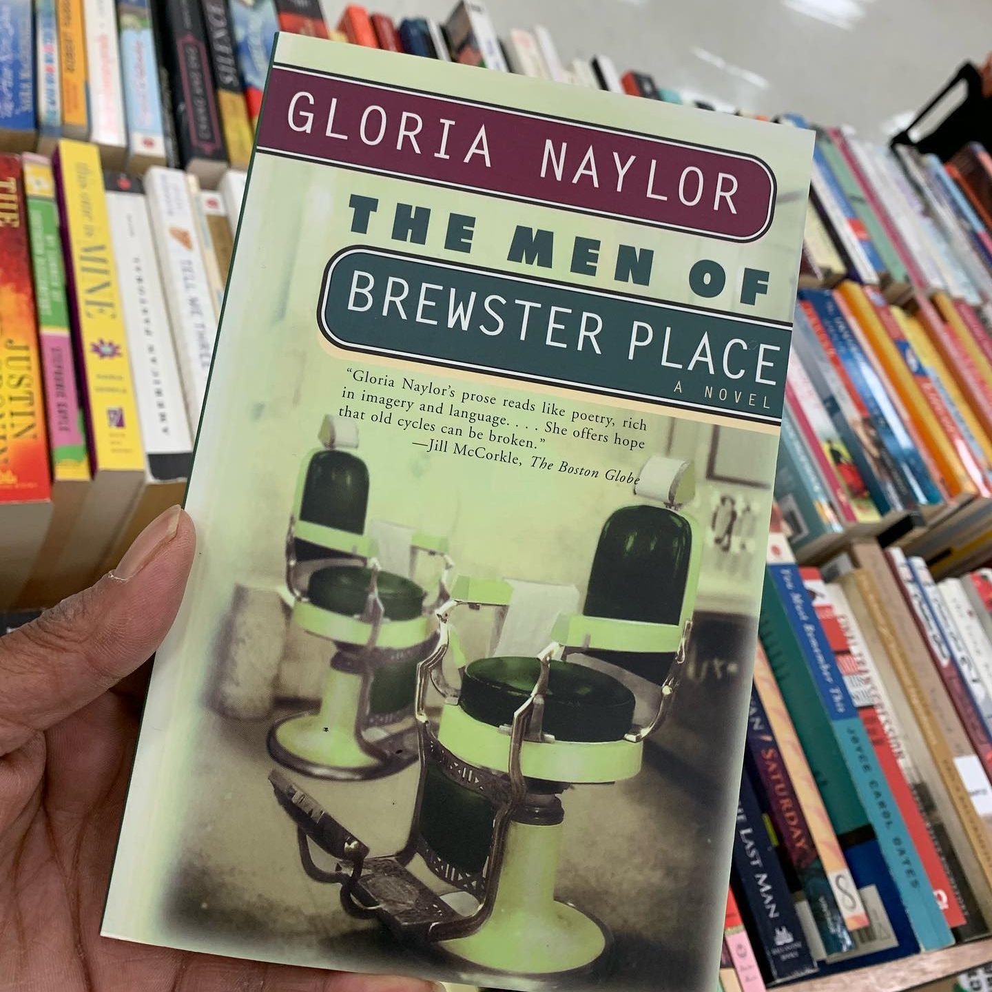 A hand holds Gloria Naylor's "The Men of Brewster Place" book, with a barbershop chair cover, in front of a shelf filled with assorted books.