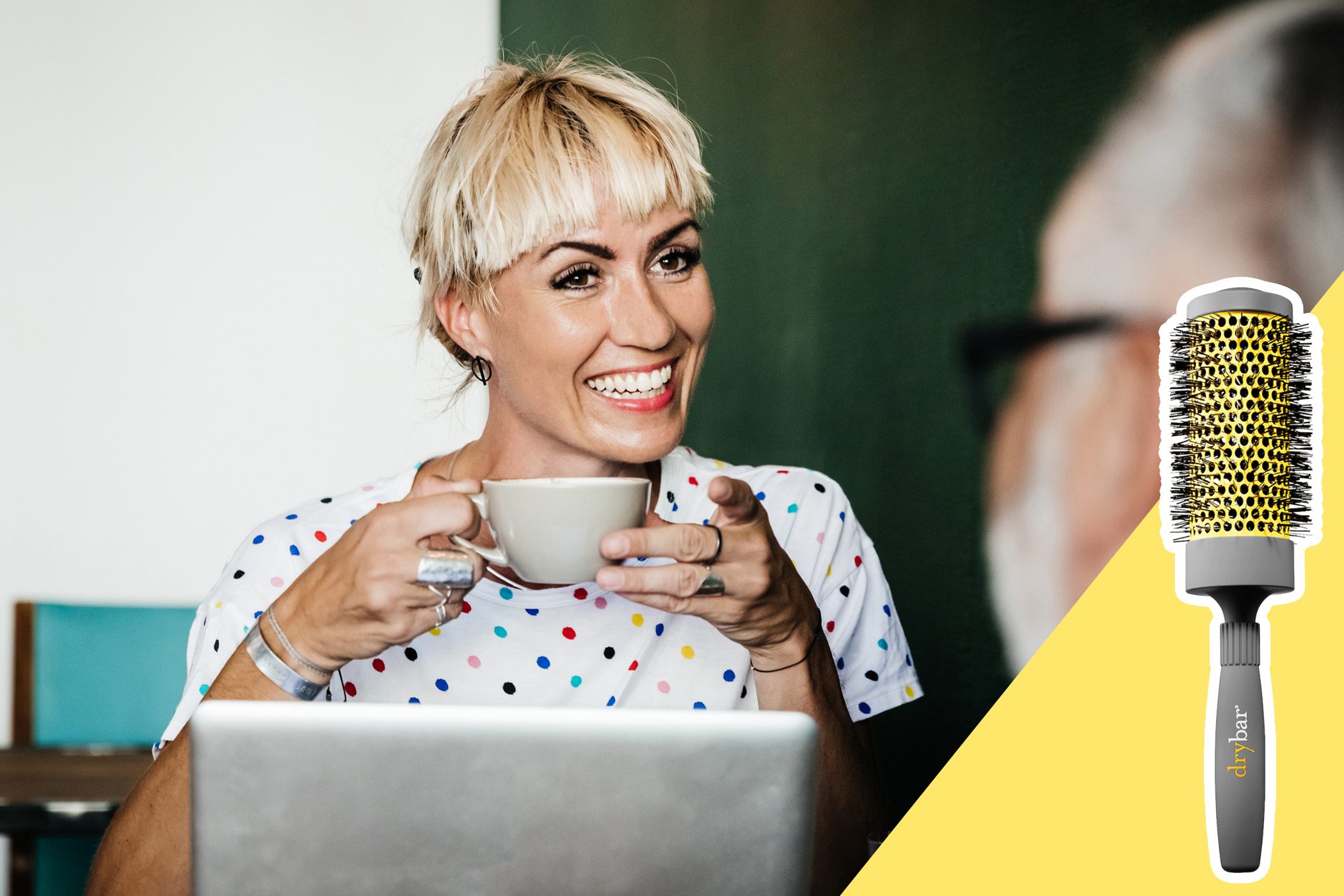 A person smiles, holding a coffee cup, in front of a laptop; Drybar hairbrush on the right over a yellow background.