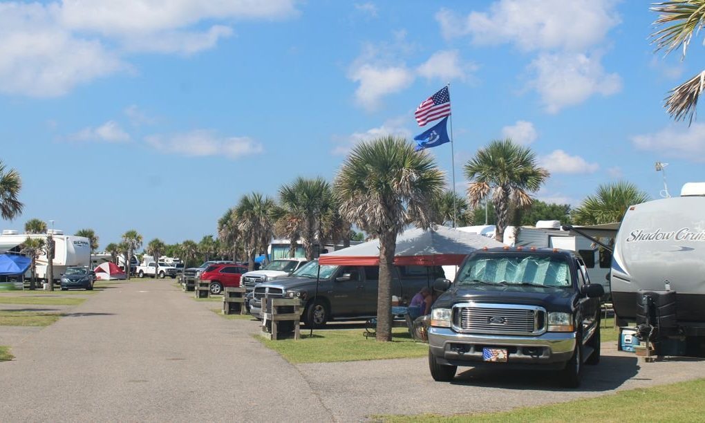 RVs parked beside a road with palm trees and flags waving, including the American flag, under a blue sky with scattered clouds.