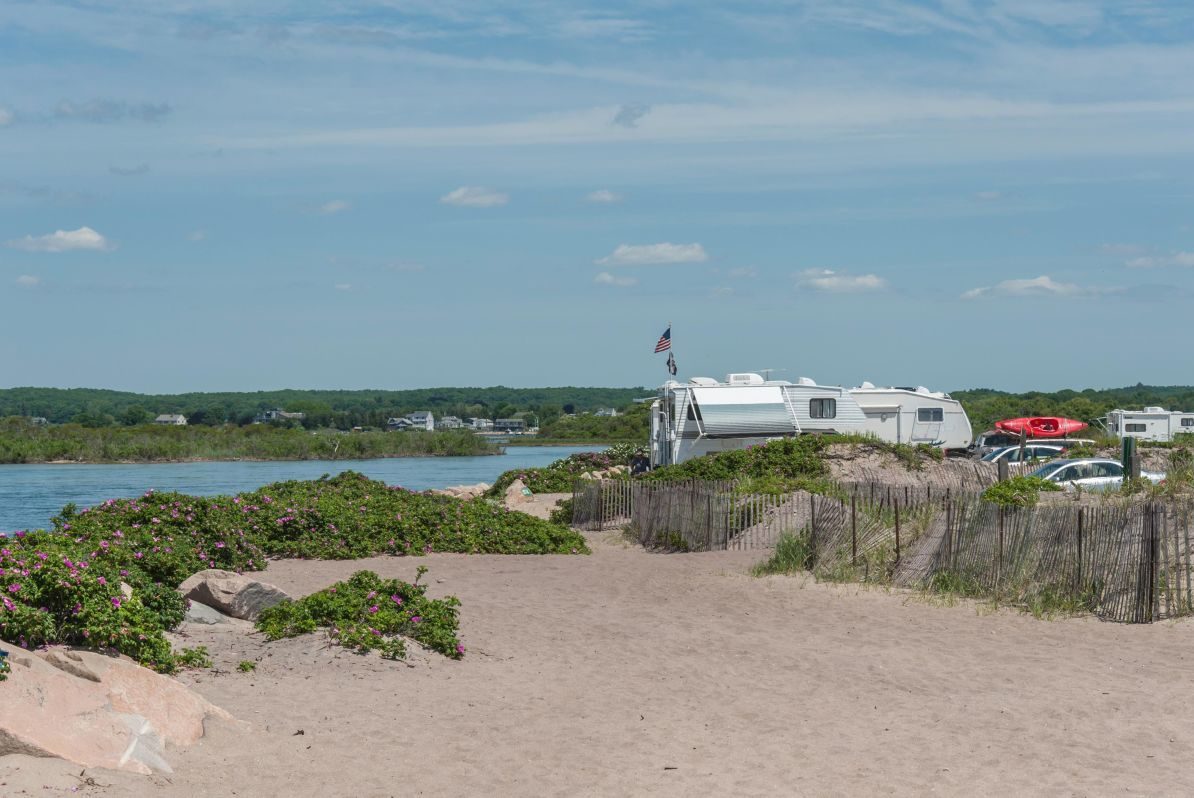 Camper parked with kayaks on sandy beach next to water, surrounded by greenery and wooden fences under a clear sky.