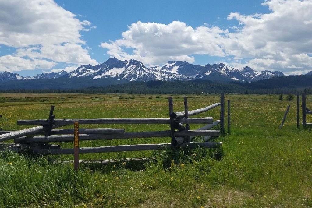 Wooden fence stands in a sunny meadow, mountains with snow caps in the background under a cloudy sky.