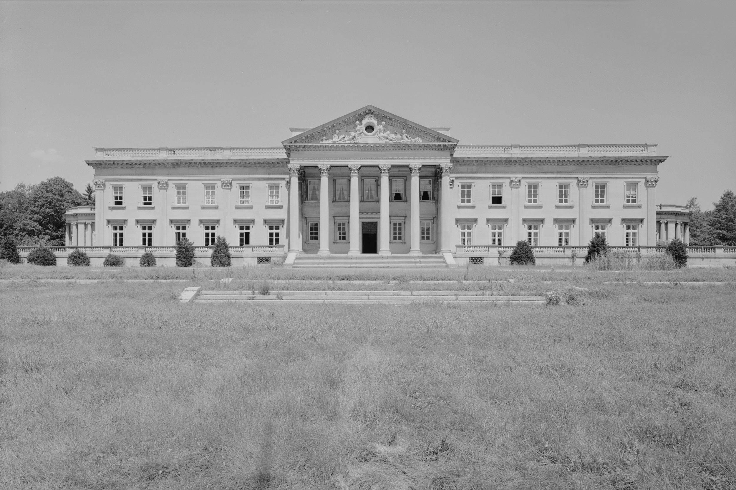 A grand, symmetrical neoclassical mansion stands in a grassy field, featuring columns, elaborate carvings, and numerous windows, amidst sparse surrounding vegetation.