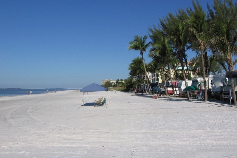 Canopy shelter stands alone; chairs underneath on a wide, sandy beach. Palm trees and buildings line the edge under a clear blue sky.