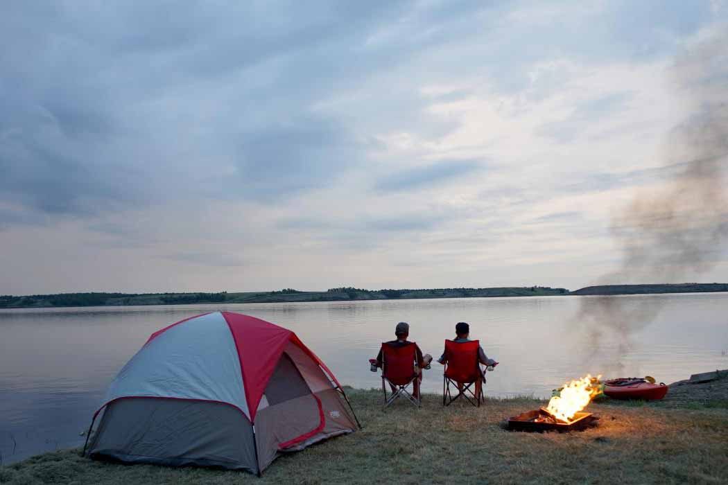 Two people sit on red chairs beside a small campfire, near a lake with a tent in the background.
