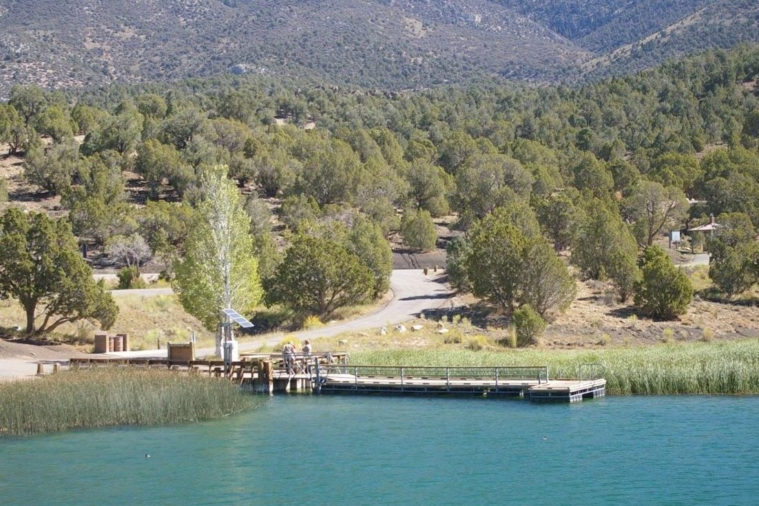 A dock extends over a turquoise lake, surrounded by green trees with mountains in the background. A small path leads to the water.