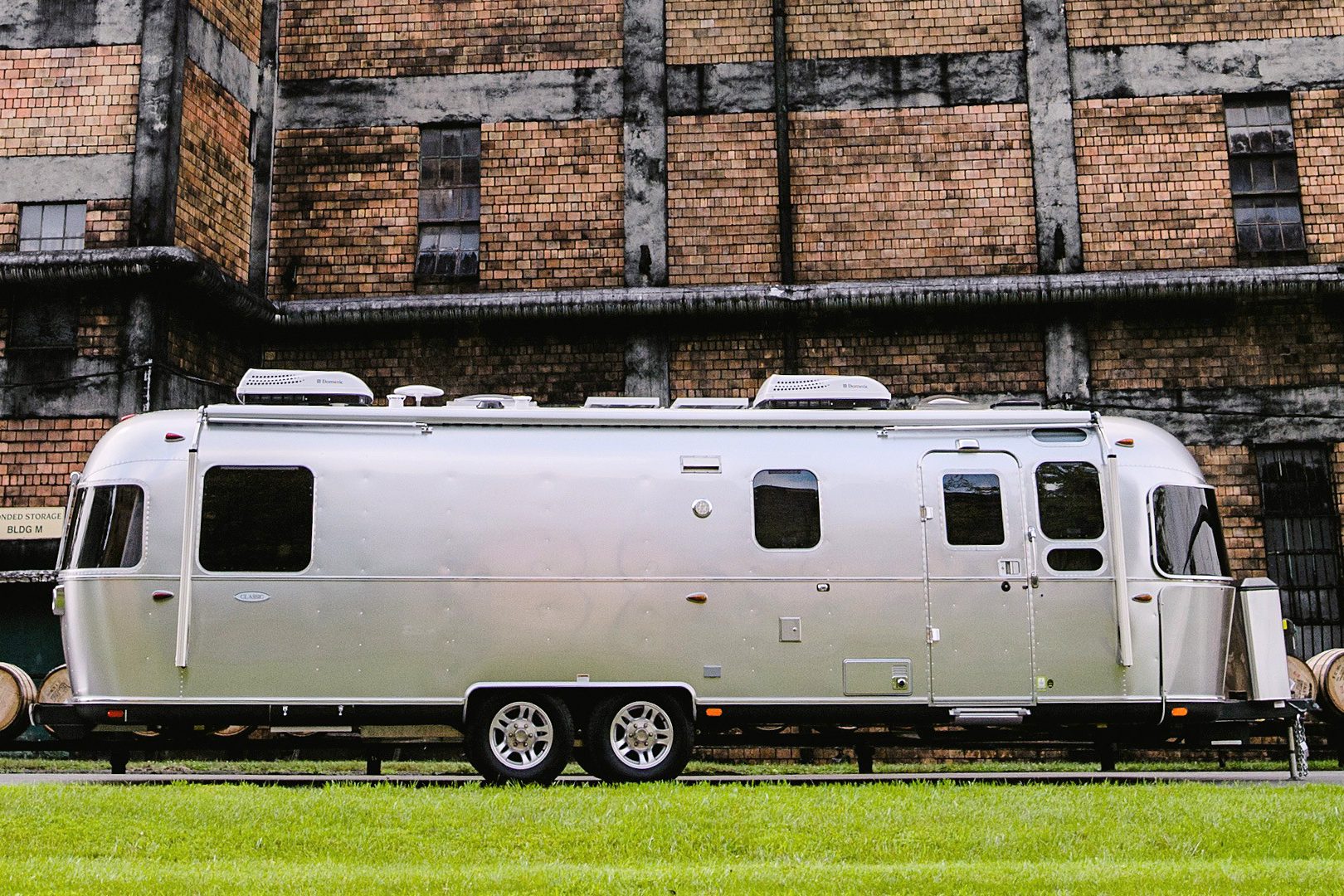 Silver travel trailer parked; background features a large brick industrial building. Grass covers the foreground, adding contrast.