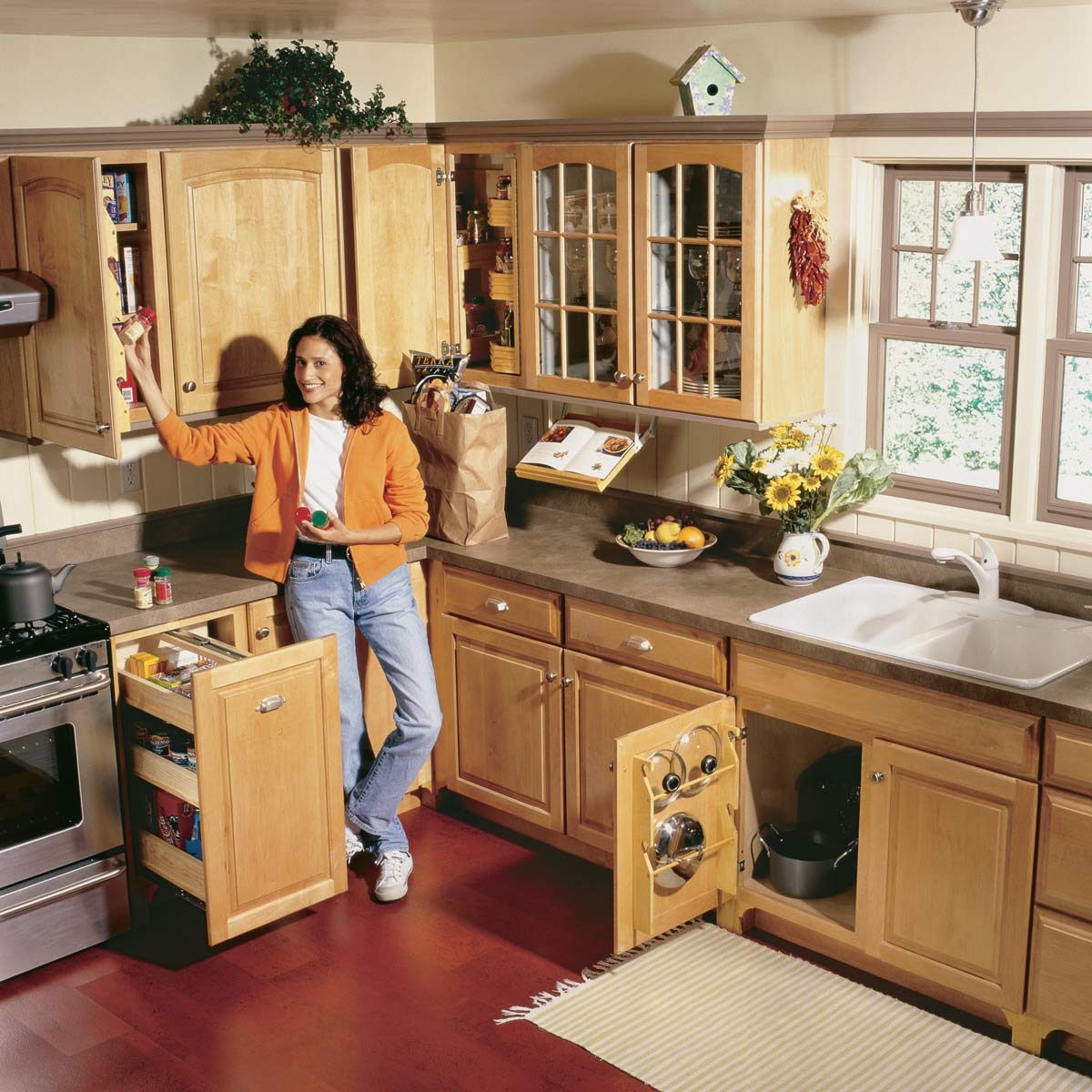 Woman reaches for spices in a kitchen with open cabinets, groceries, large window, and sunflowers on the countertop near the sink.