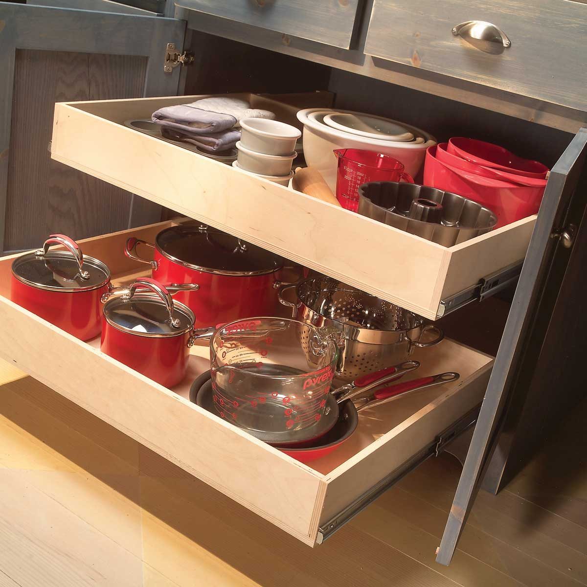 Wooden drawers slide open, revealing red cookware and glass measuring cups, organized in a kitchen cabinet.