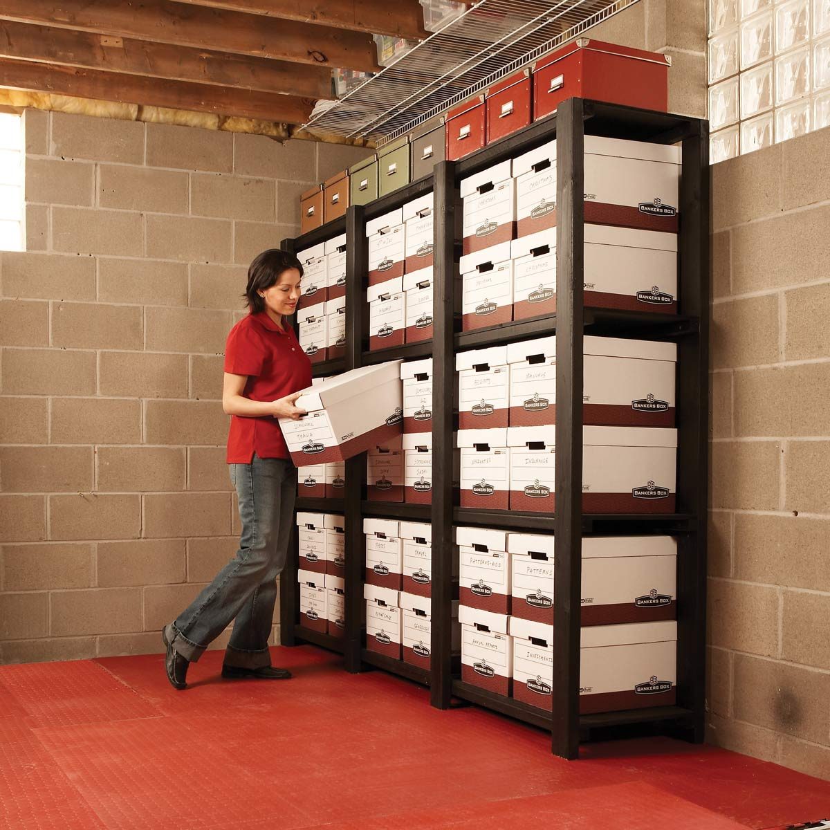 Person organizing cardboard boxes on shelves in a basement with concrete walls and a red floor.
