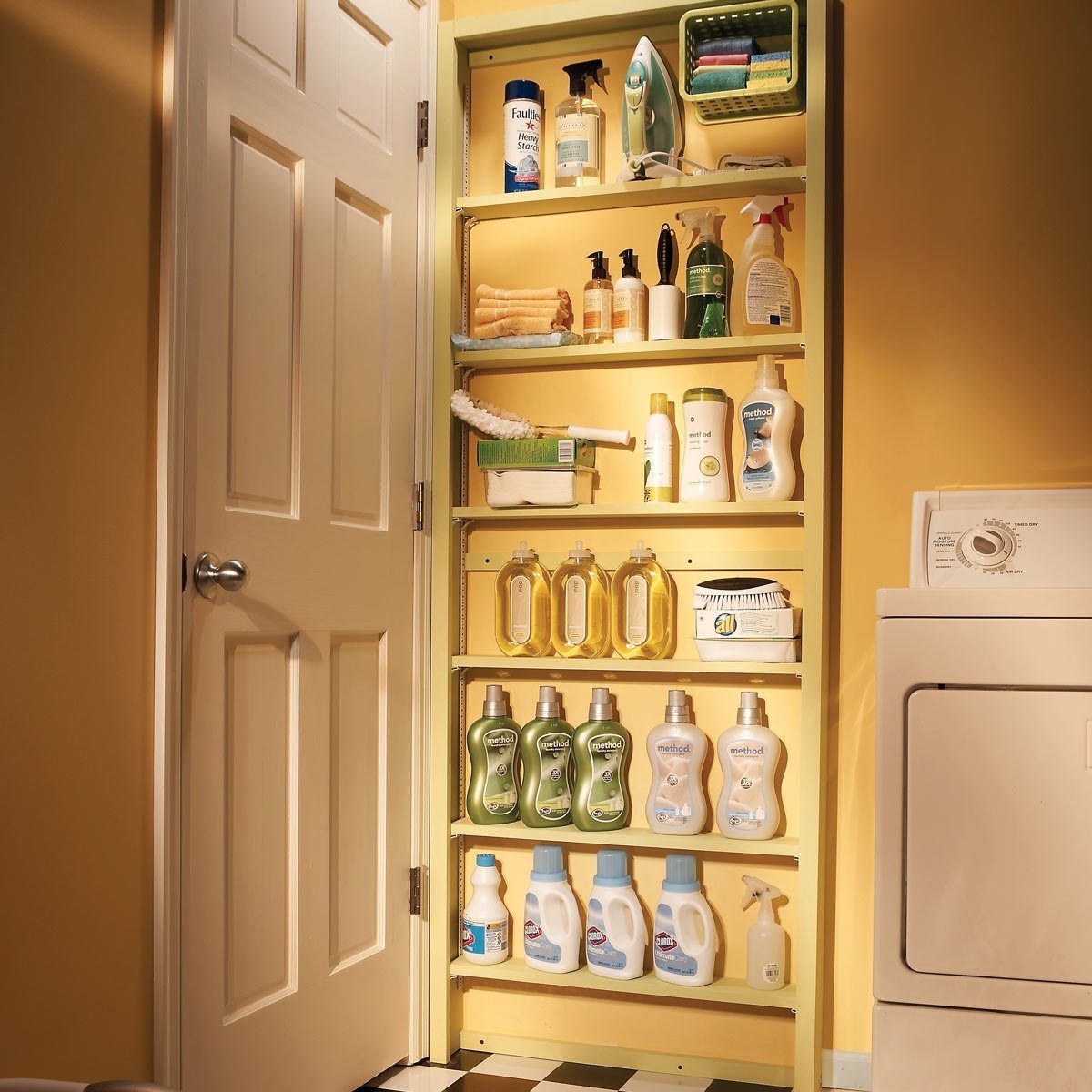 Shelves hold various cleaning supplies, organized neatly beside a closed dryer in a warm, yellow-lit laundry room.