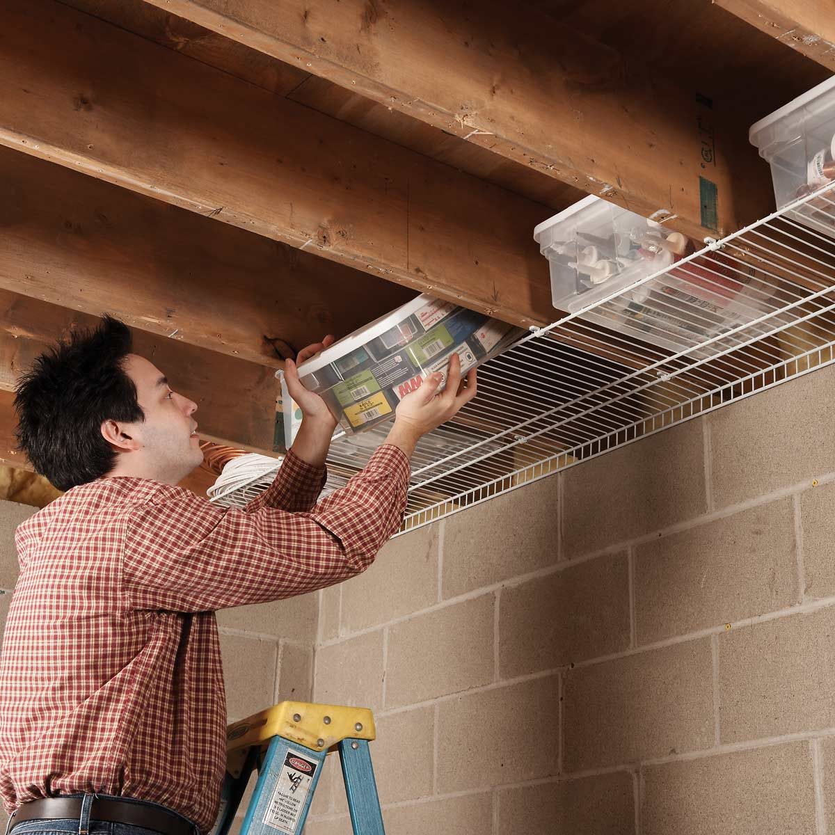 Person places a plastic storage bin on a wire shelf overhead, standing on a ladder in a basement with exposed beams and cinder block walls.