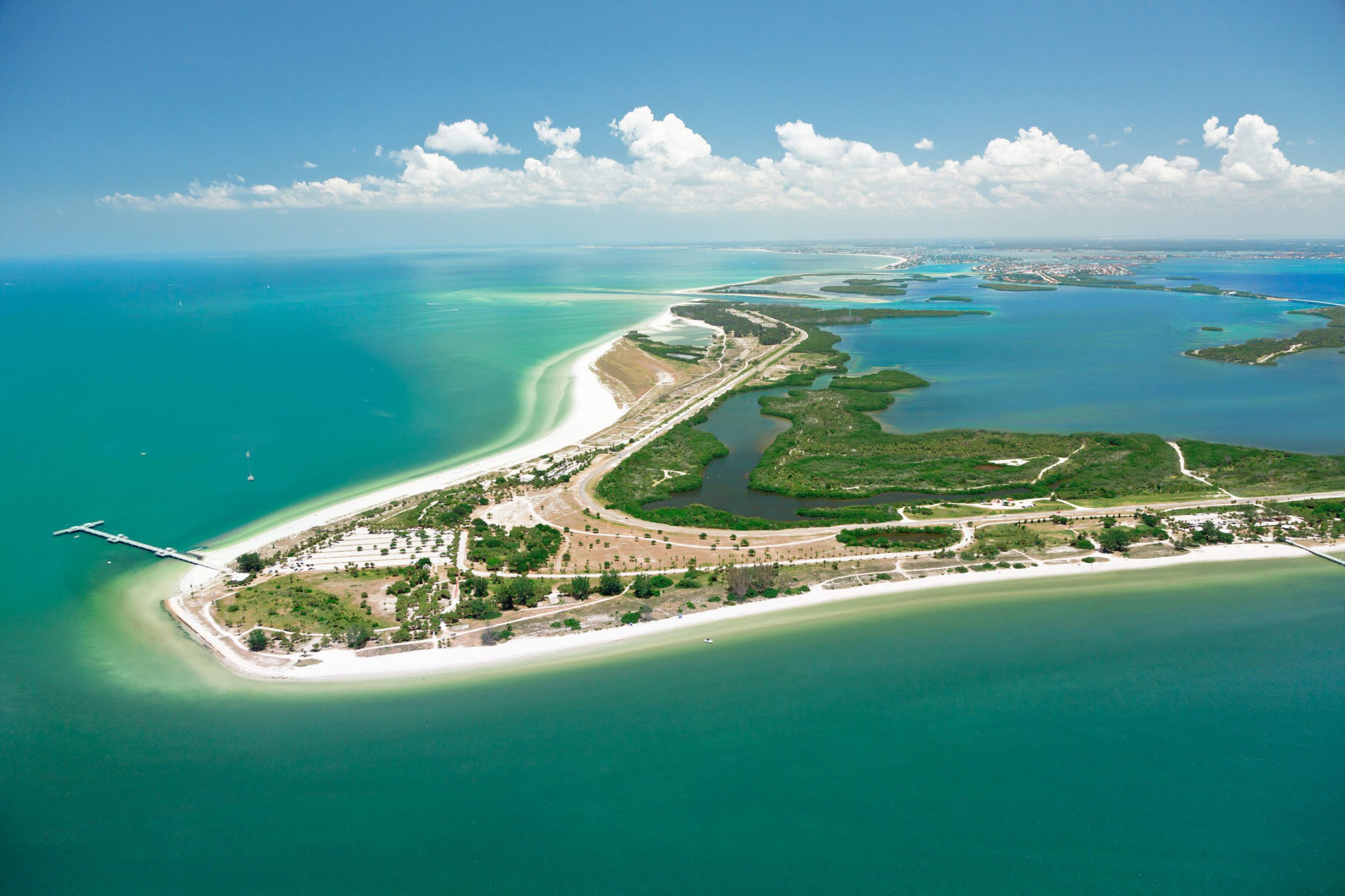 Island stretches into turquoise ocean, bordered by white sandy beaches, with lush greenery and a pier extending into the water, under a clear blue sky.