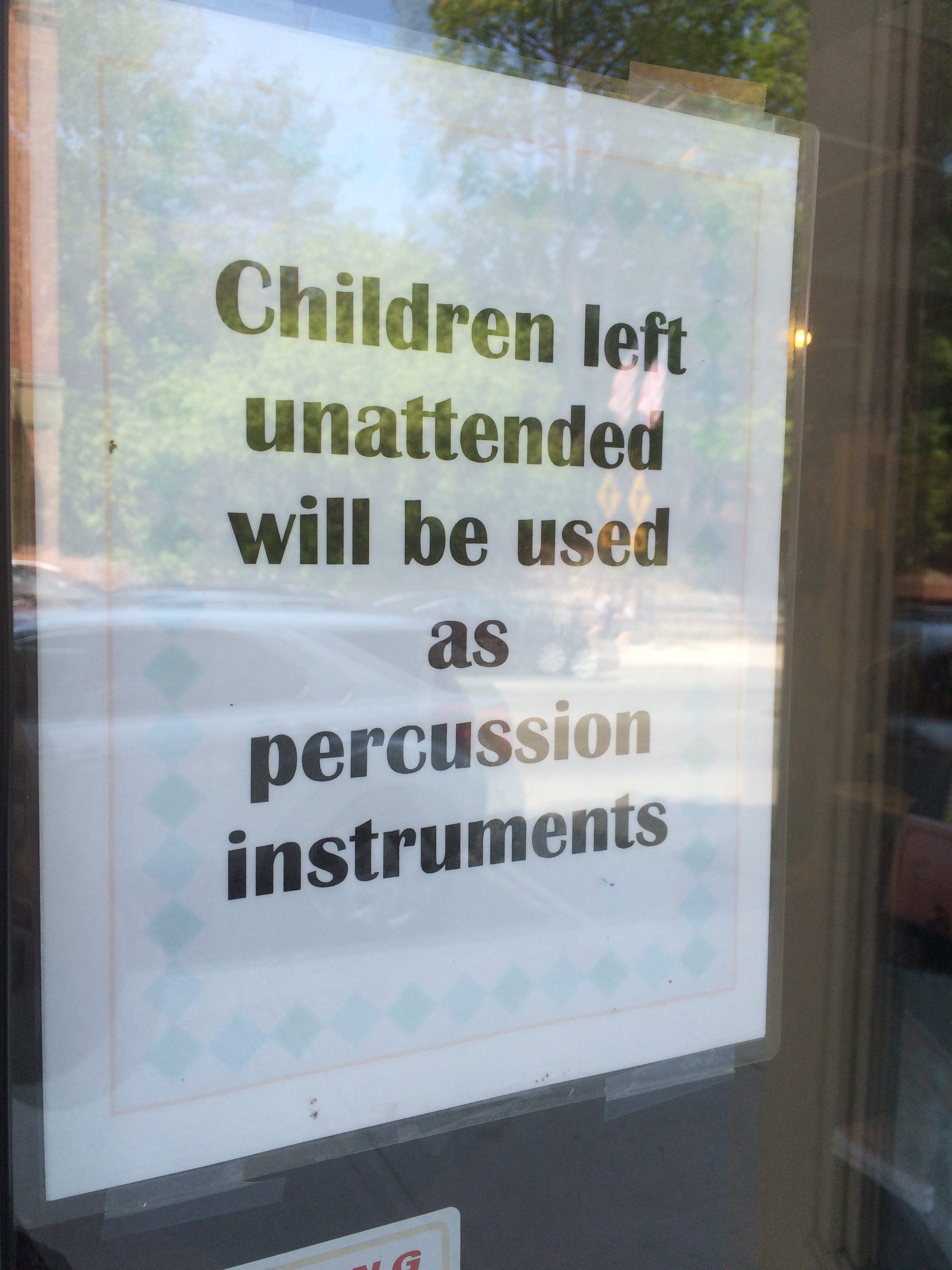 Sign displays humorous message: "Children left unattended will be used as percussion instruments." Positioned on glass with trees and cars reflected.