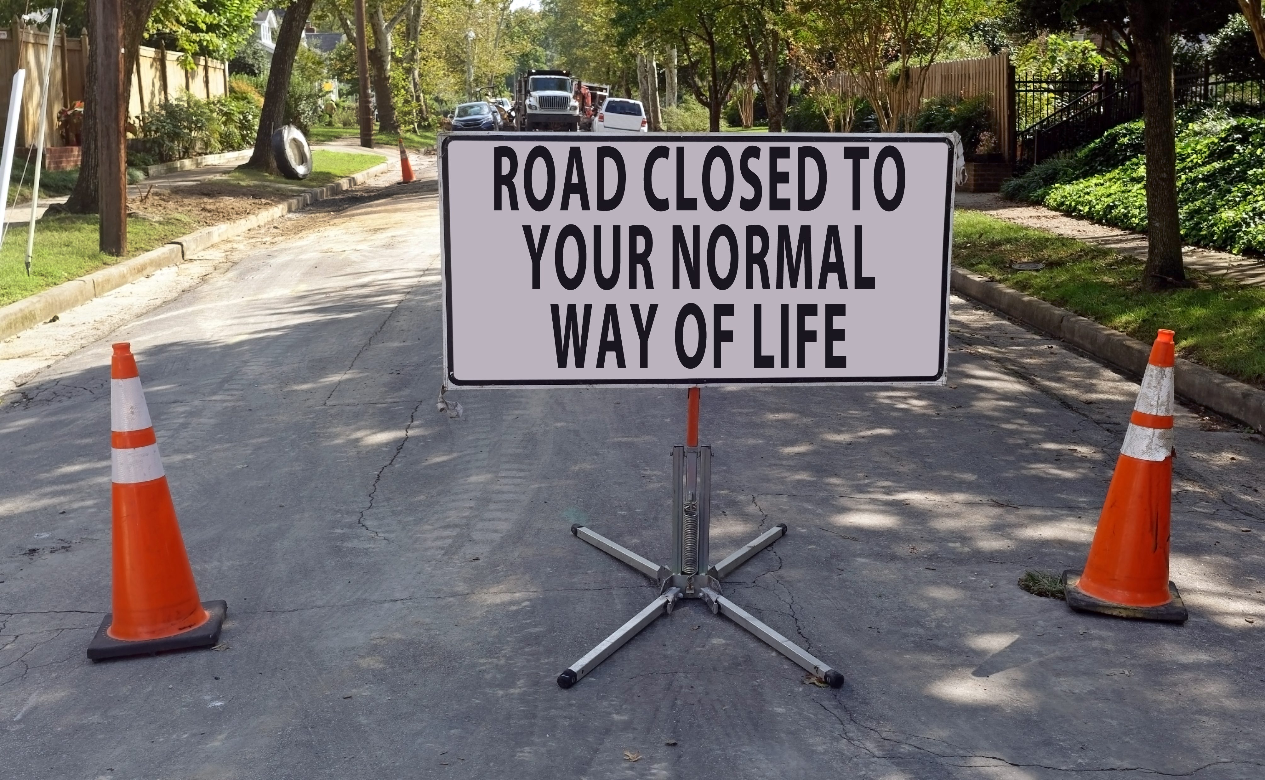 Sign displays "ROAD CLOSED TO YOUR NORMAL WAY OF LIFE," placed on a street between orange cones with trees and vehicles in the background.