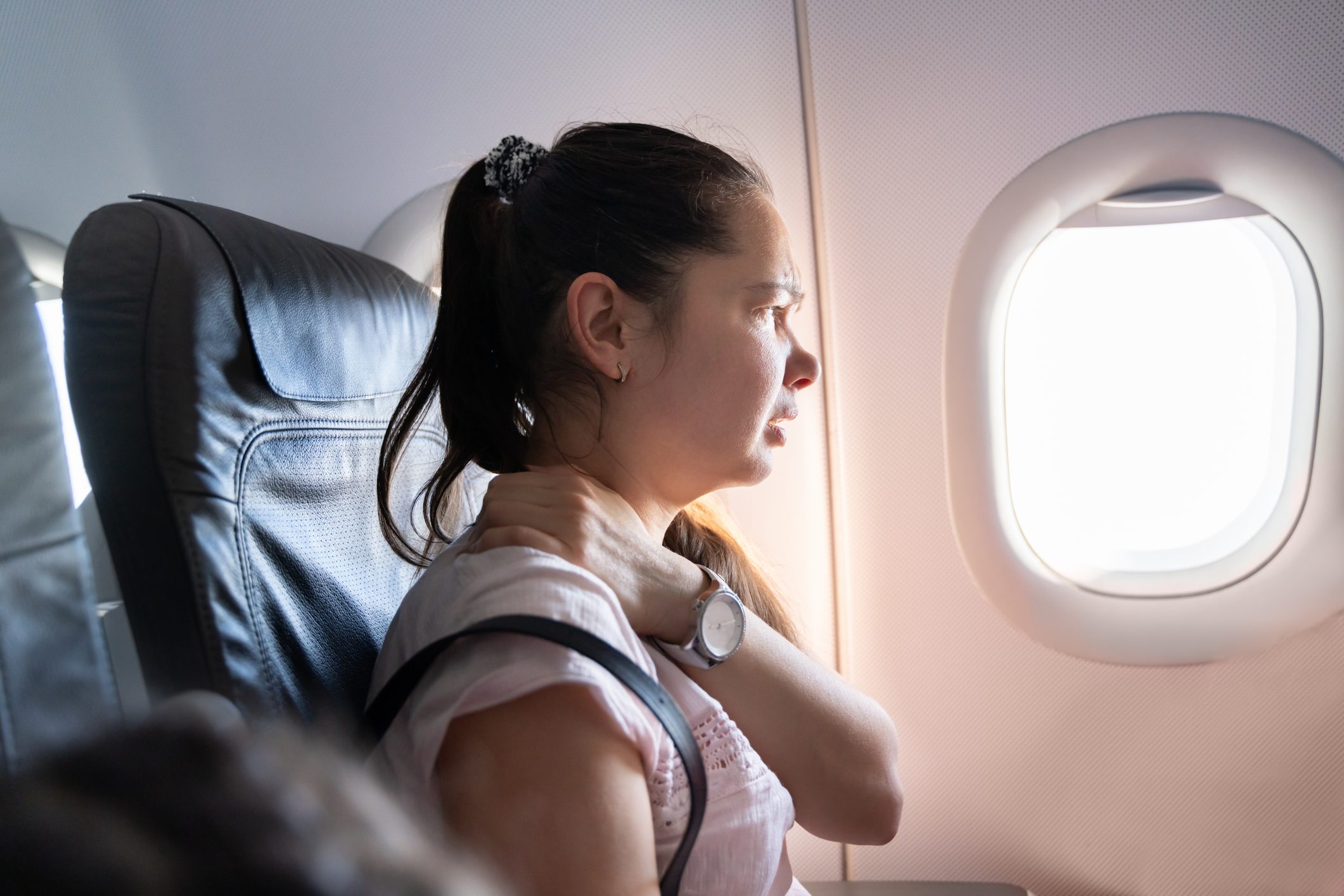 Woman holding neck, seated, looking out airplane window.