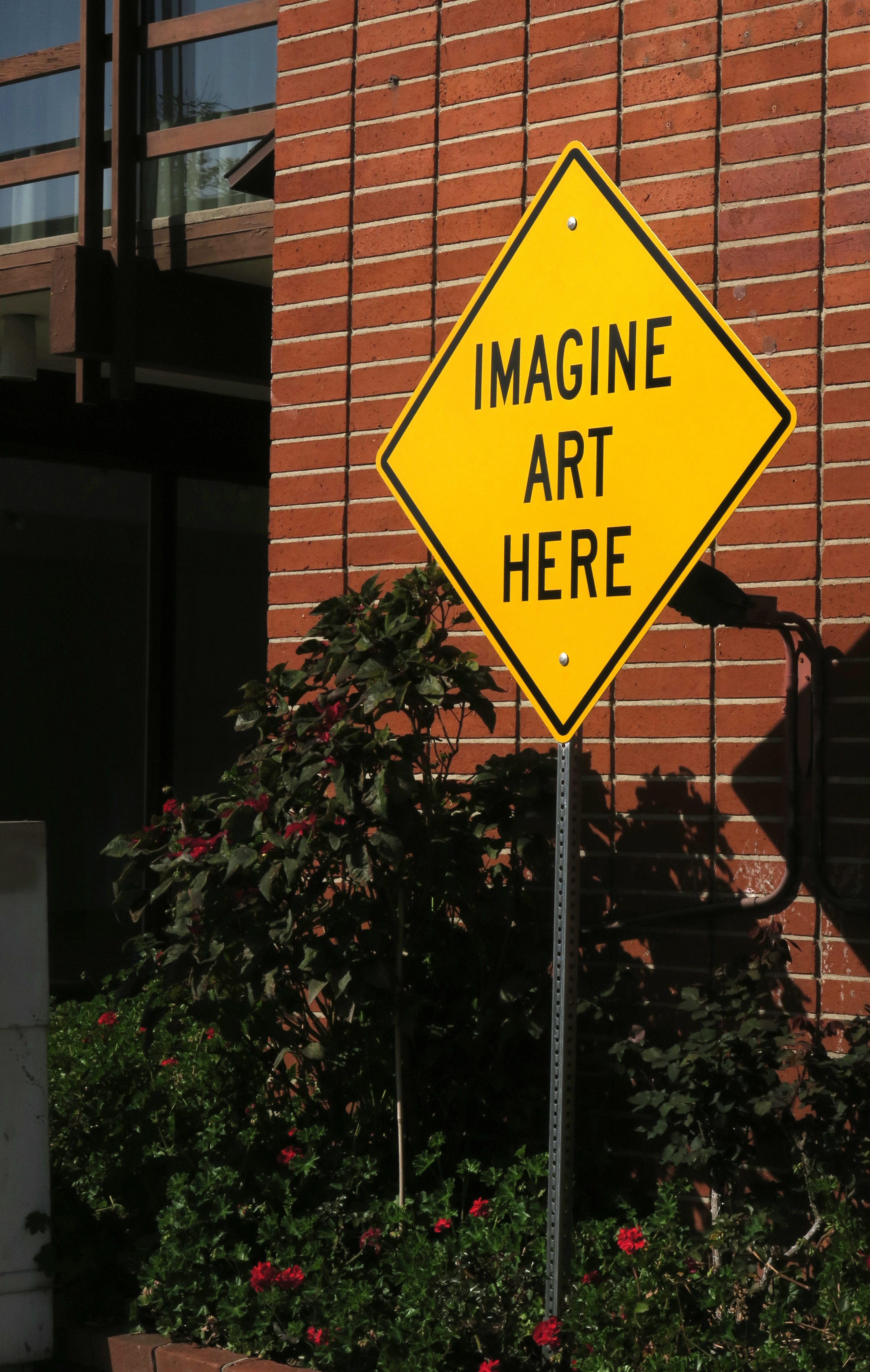 A yellow sign reads "IMAGINE ART HERE" beside a brick wall, surrounded by green foliage and red flowers.