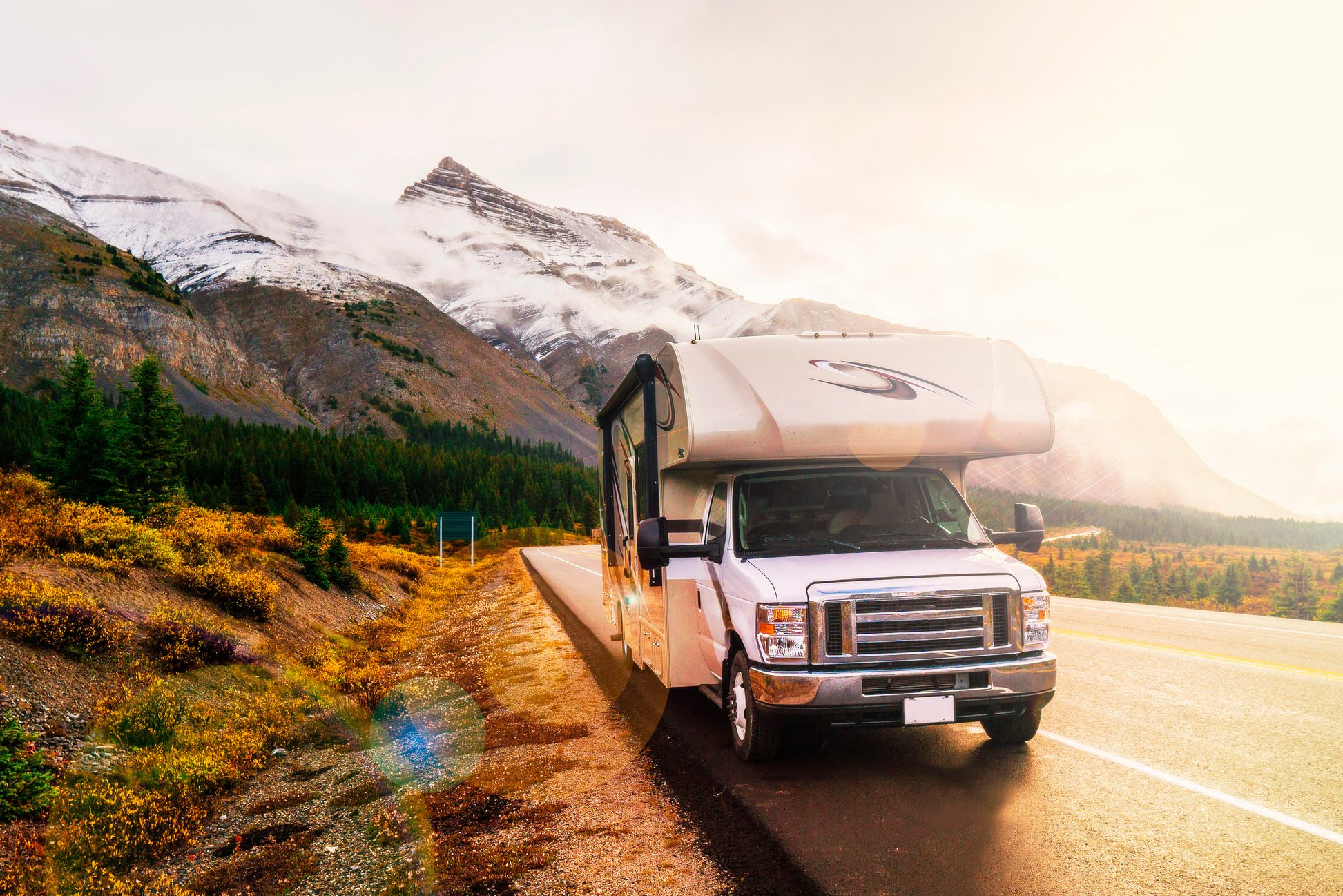 RV parked along a scenic mountain road, with autumn foliage and snow-capped peaks in the background under a bright sky.