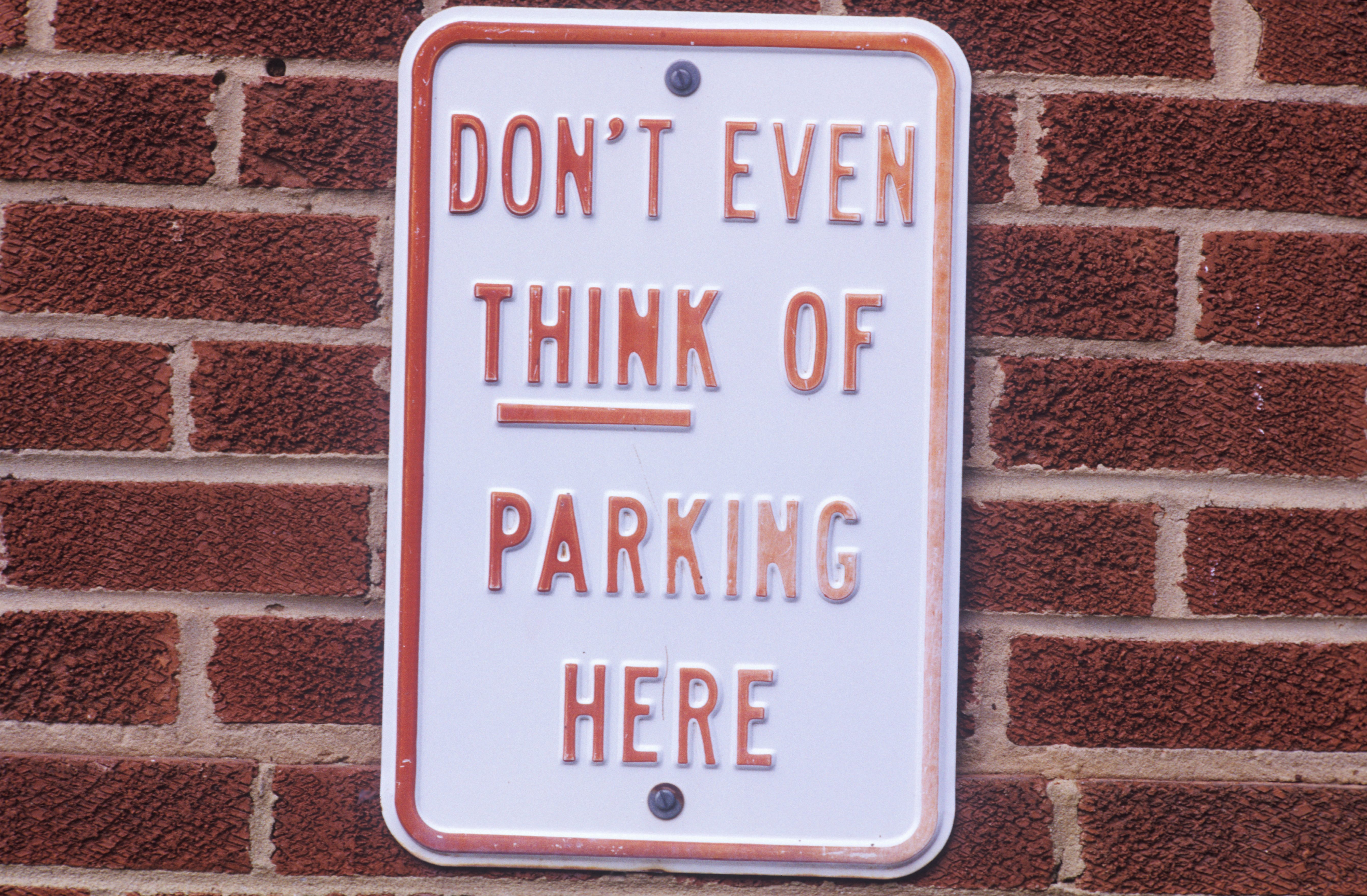 A sign reads "DON'T EVEN THINK OF PARKING HERE," mounted on a red brick wall.