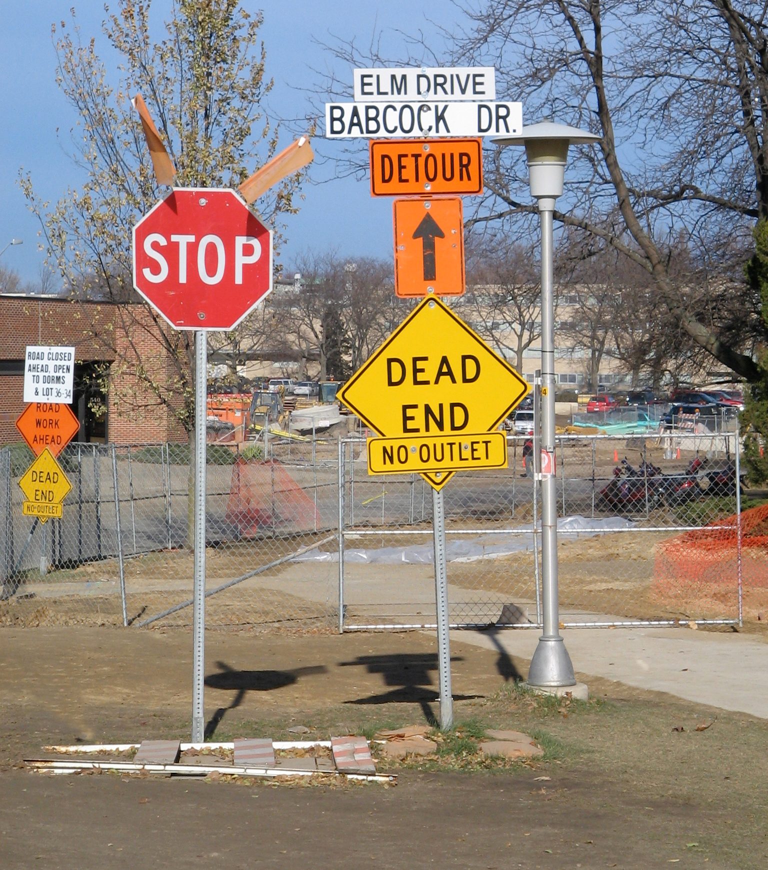 Signs display "STOP," "DEAD END," and "DETOUR," situated near a construction site with fencing and trees, indicating road changes ahead.