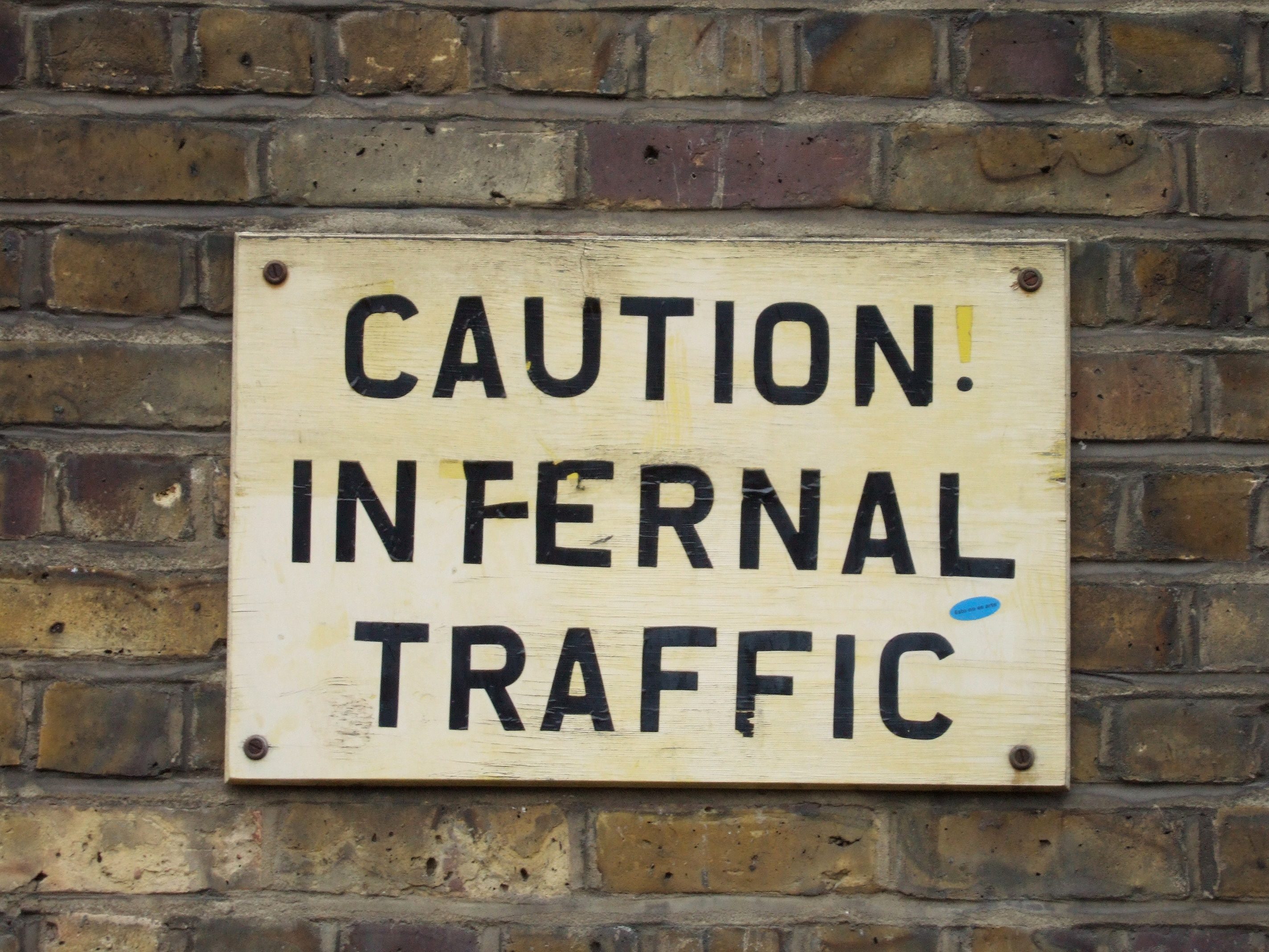 Sign warns "CAUTION! INFERNAL TRAFFIC," mounted on a brick wall.