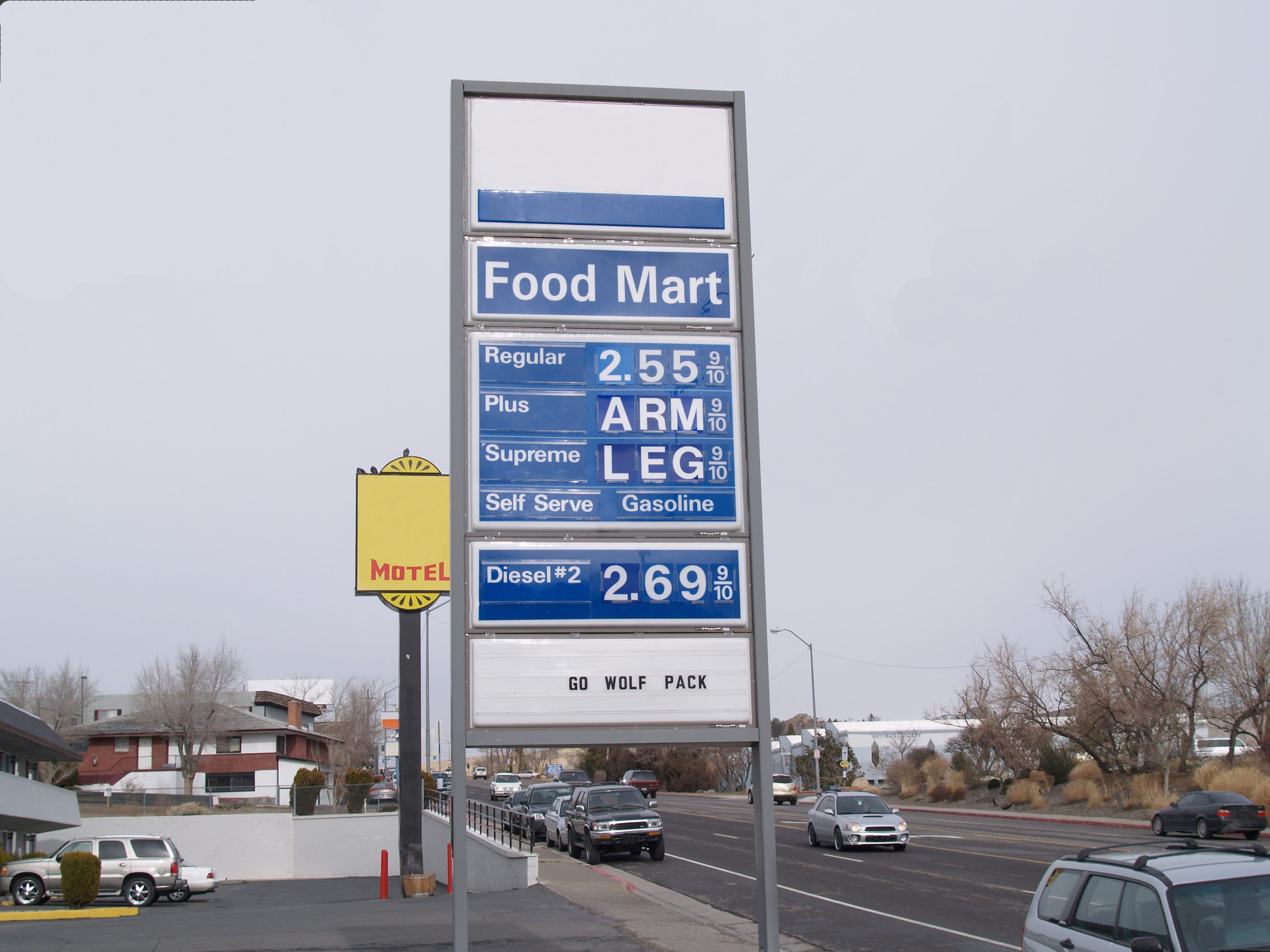 Sign displays fuel prices humorously as "ARM" and "LEG" beside regular prices, set in a suburban street with cars and trees.