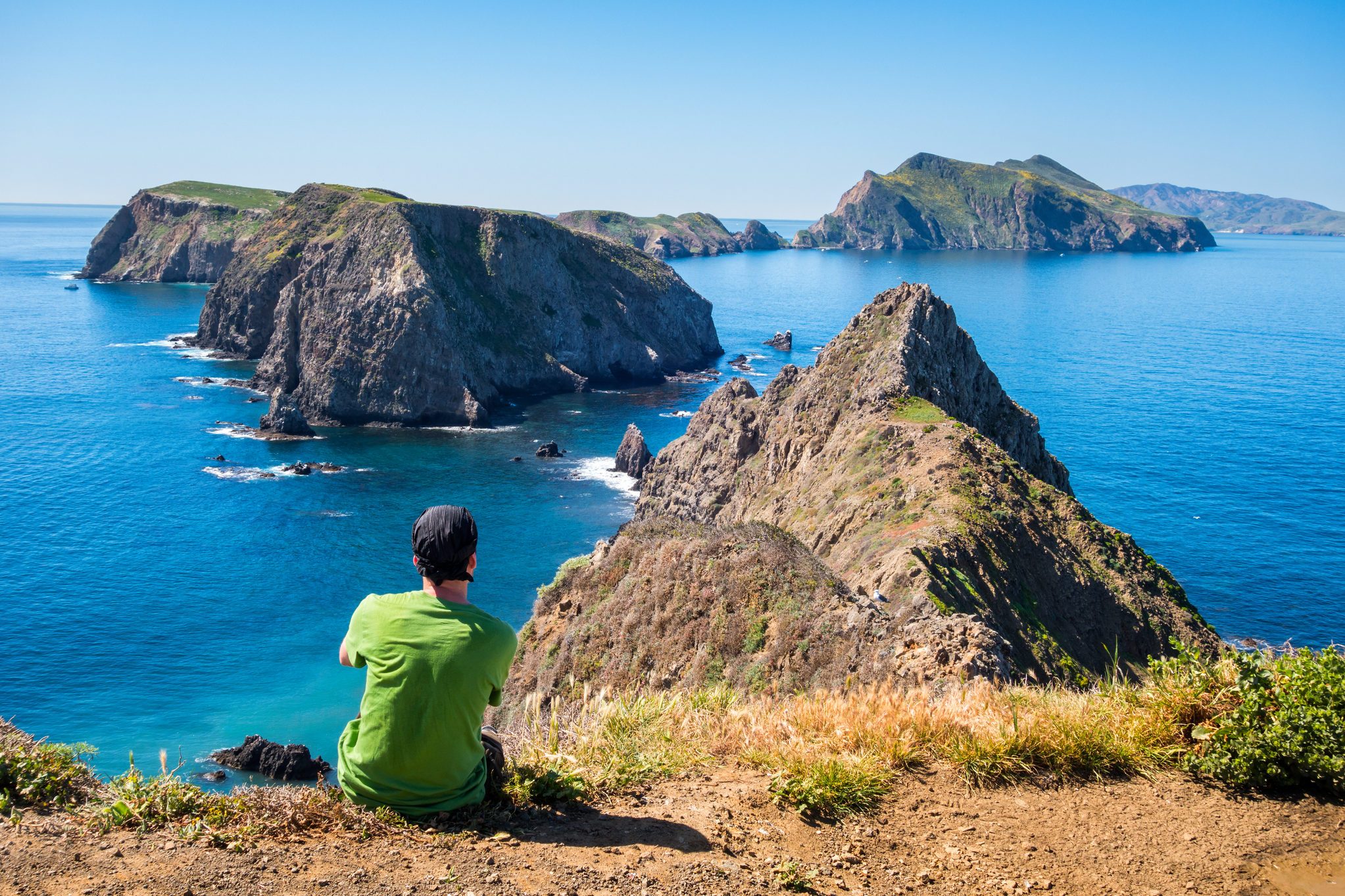 Person sits on cliff edge overlooking rocky islands and vibrant blue ocean under clear sky.